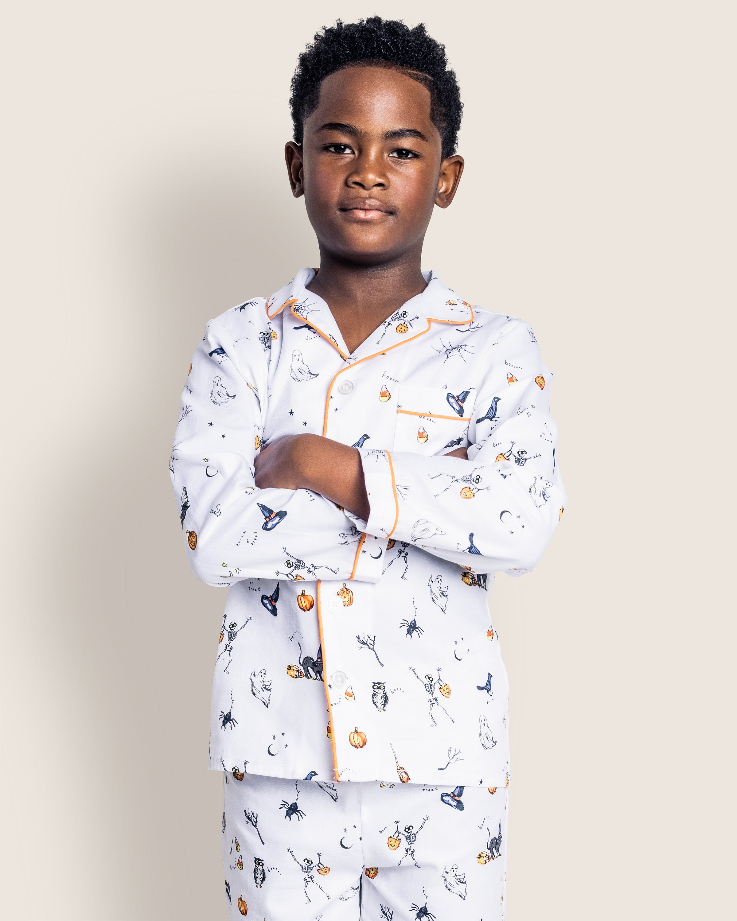 A child with curly hair stands confidently, arms crossed, in a luxurious Kids Twill Pajama Set in Trick or Treat by Petite Plume. Adorned with pumpkins, ghosts, and bats on a neutral beige background, the playful design highlights their sleepwear.