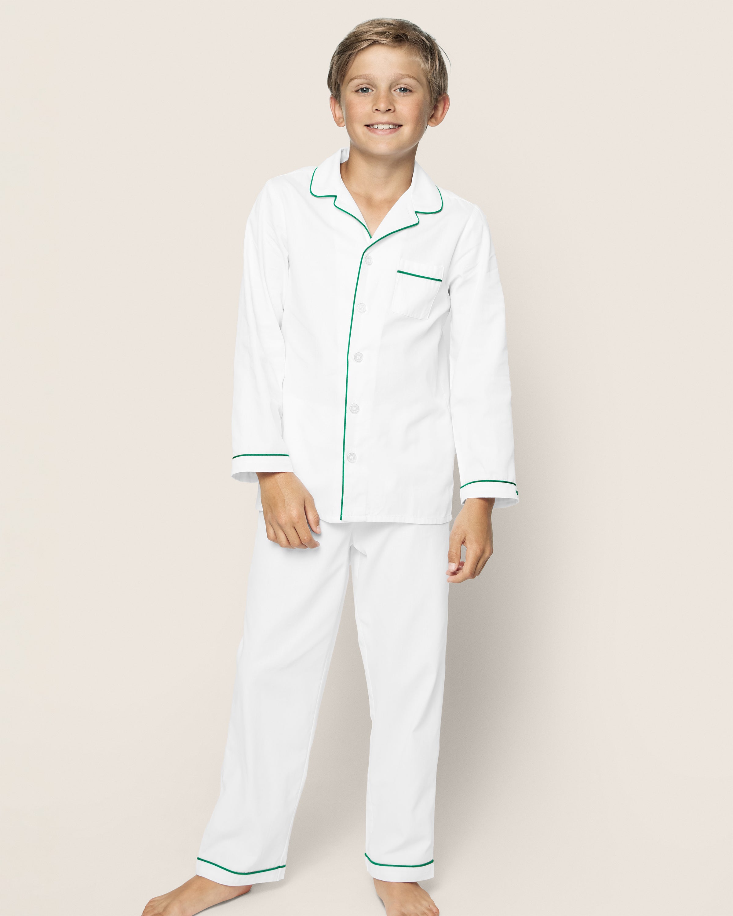 A smiling boy stands barefoot on a beige background, wearing Petite Plumes cozy Kids Twill Pajama Set in white with green piping. The button-up shirt includes a pocket and is paired with matching pants.