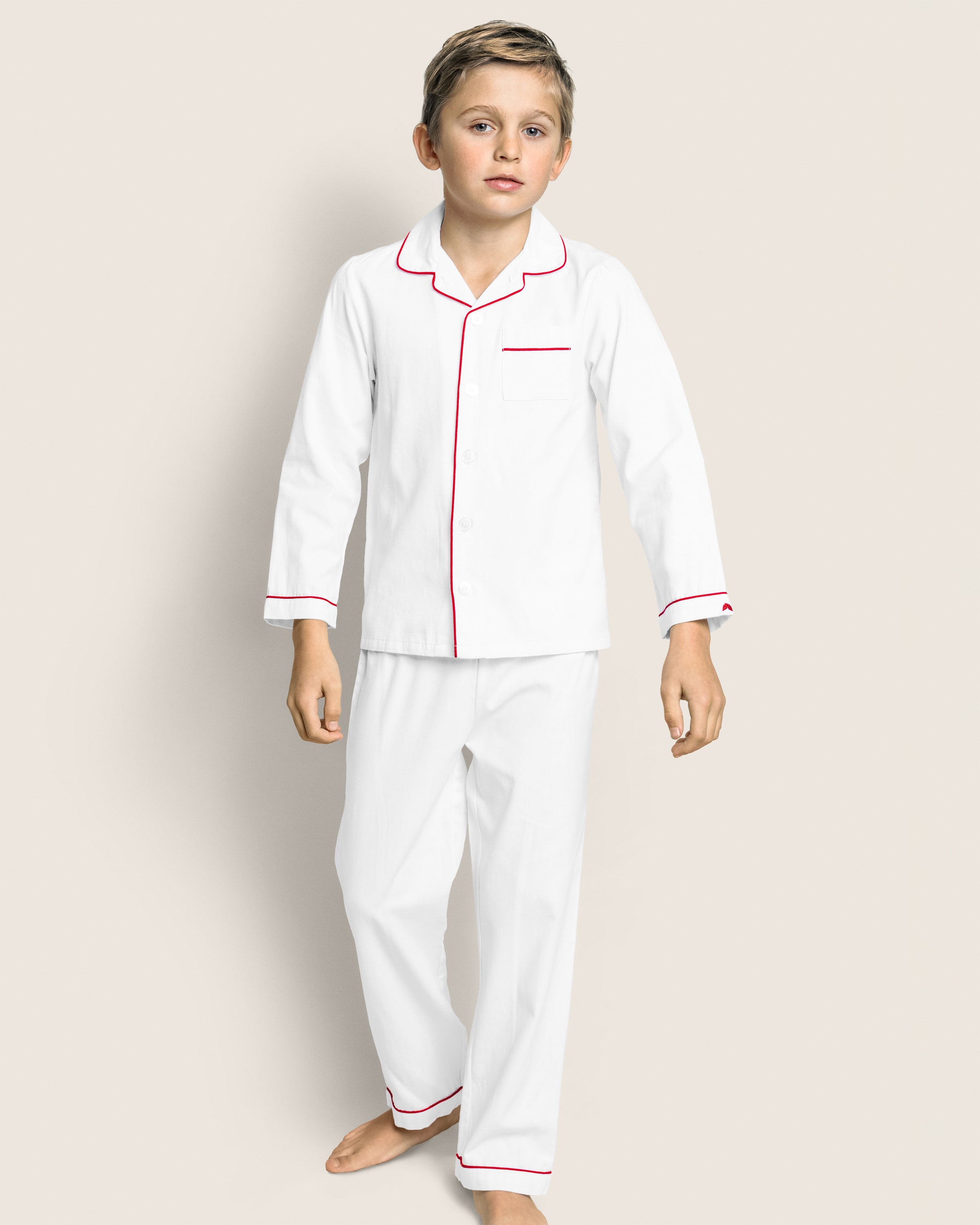 A child wears Petite Plumes Kids Twill Pajamas in white with red piping, made from flame-retardant cotton. The set includes a long-sleeved button-up top with a pocket and matching pants. The child stands barefoot against a plain, light background.