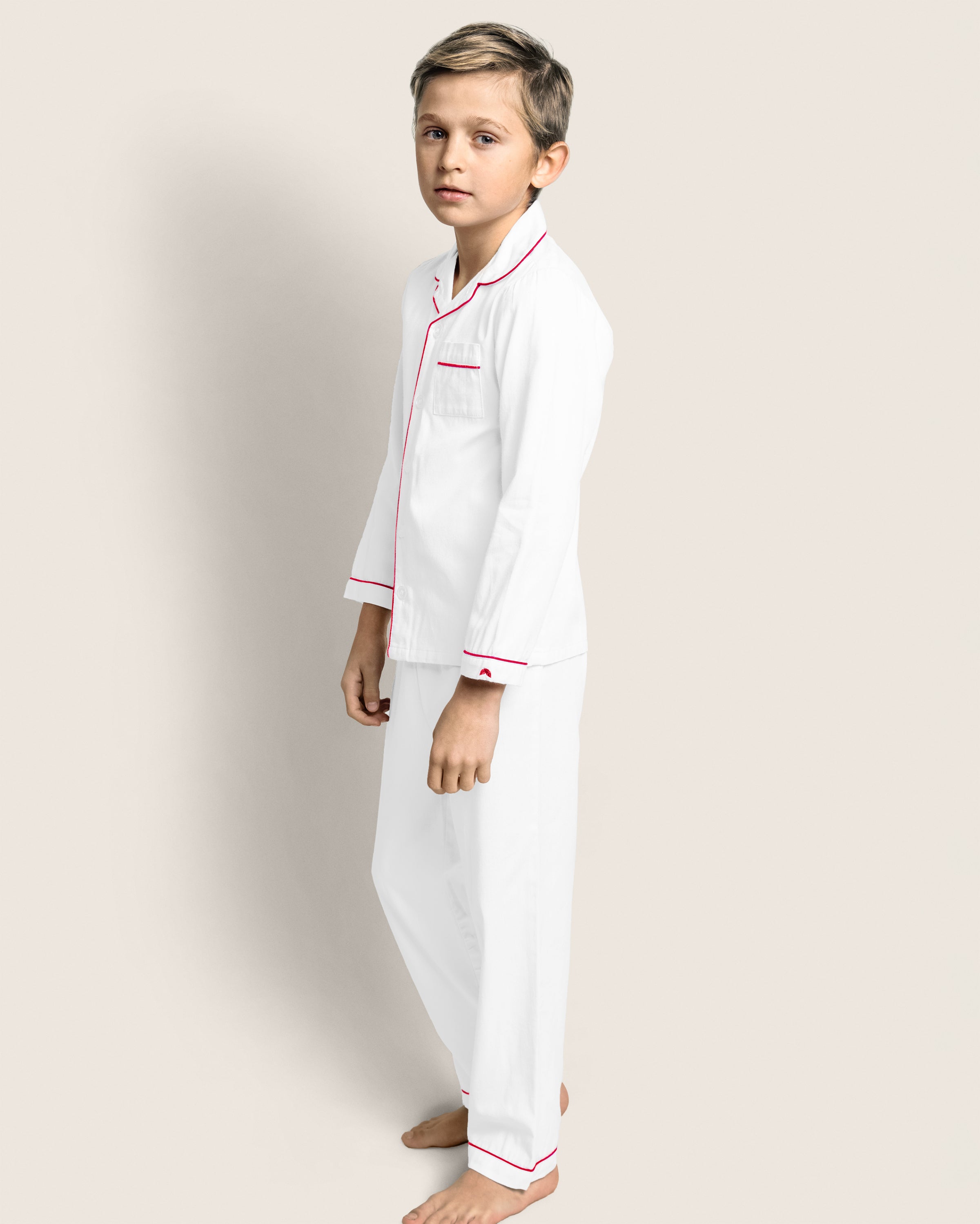 A child with short hair stands barefoot, wearing Petite Plumes Kids Twill Pajamas in White with Red Piping. The flame retardant cotton set features long sleeves and pants, offering comfort and safety as the child gazes towards the camera against a plain light background.