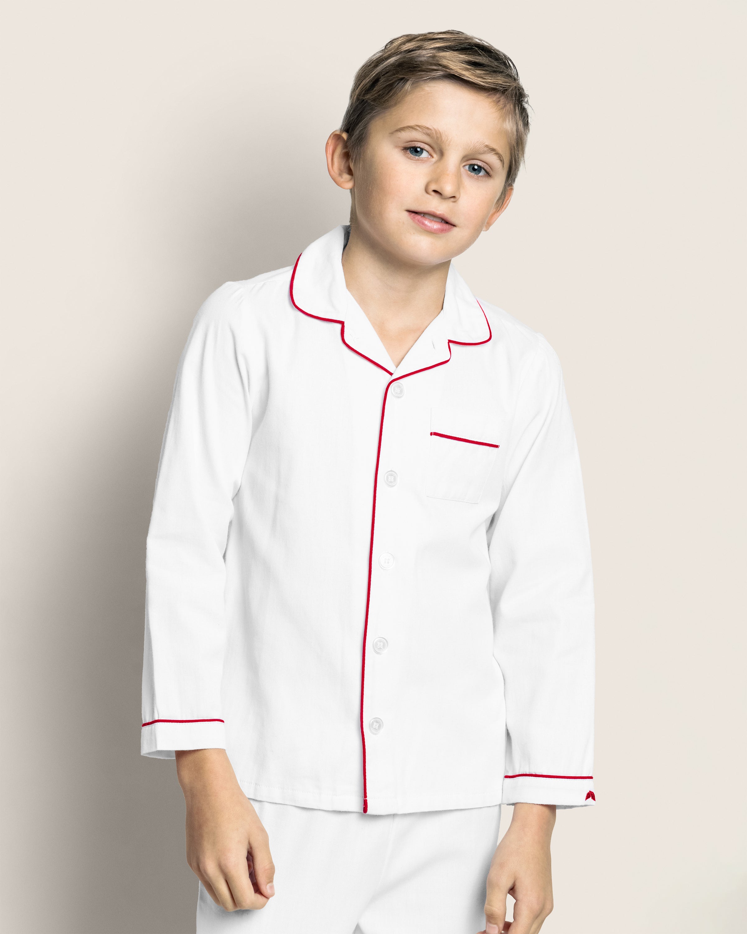 A young boy stands against a light background wearing Petite Plumes Kids Twill Pajamas in White with Red Piping, crafted from flame-retardant cotton, complementing his slight smile perfectly.