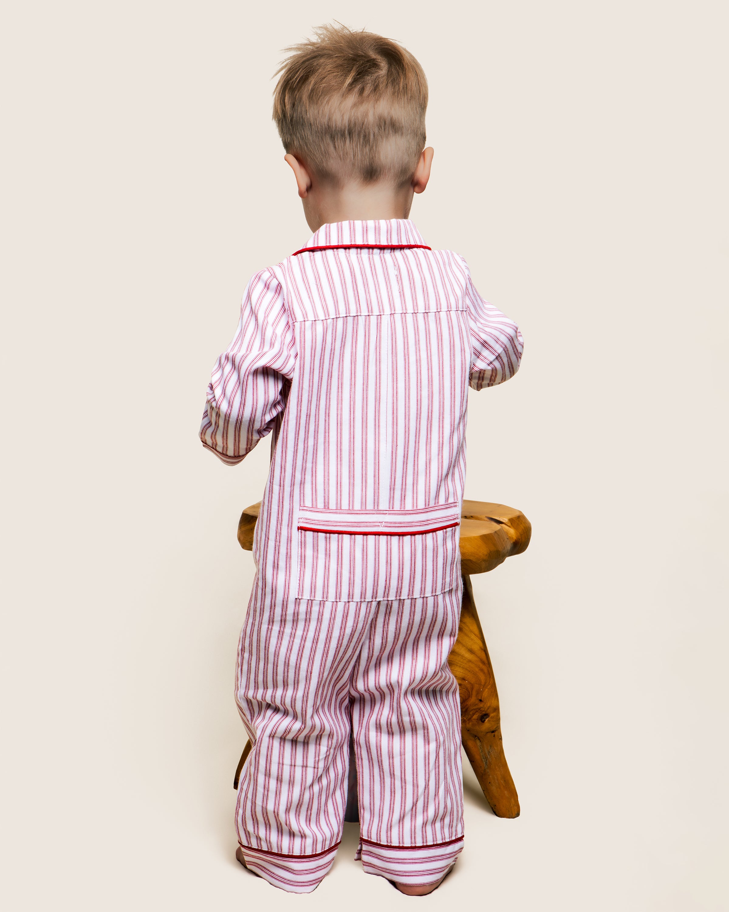 A toddler with short blond hair stands facing away, wearing Petite Plumes Babys Twill Romper in Antique Red Ticking. The child leans on a small, wooden, round stool against a plain background.