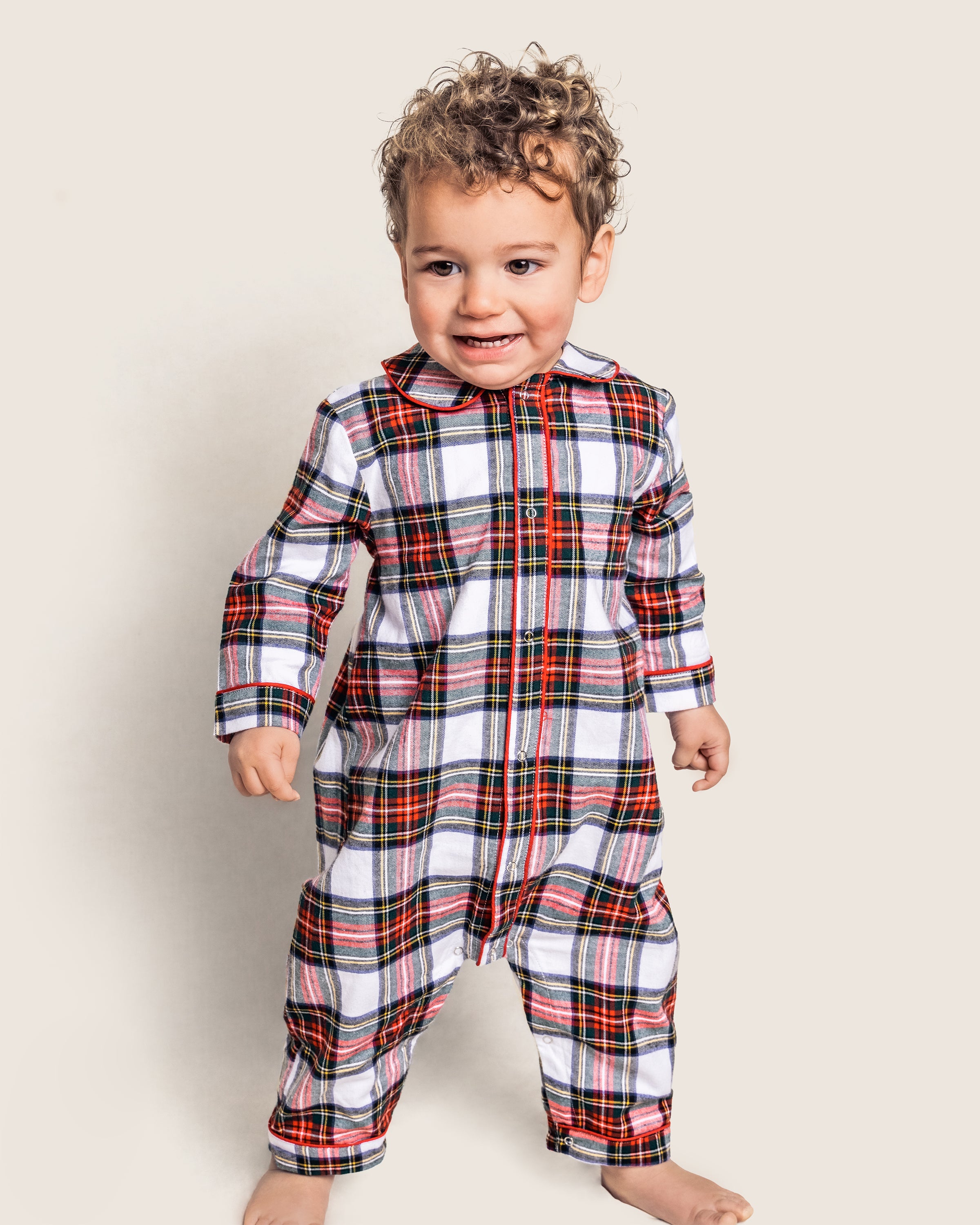 A toddler stands barefoot against a neutral backdrop, smiling and looking to the side, wearing Petite Plumes Babys Brushed Cotton Cambridge Romper in Balmoral Tartan, featuring a yarn-dyed red, white, and black pattern inspired by Scottish tradition.
