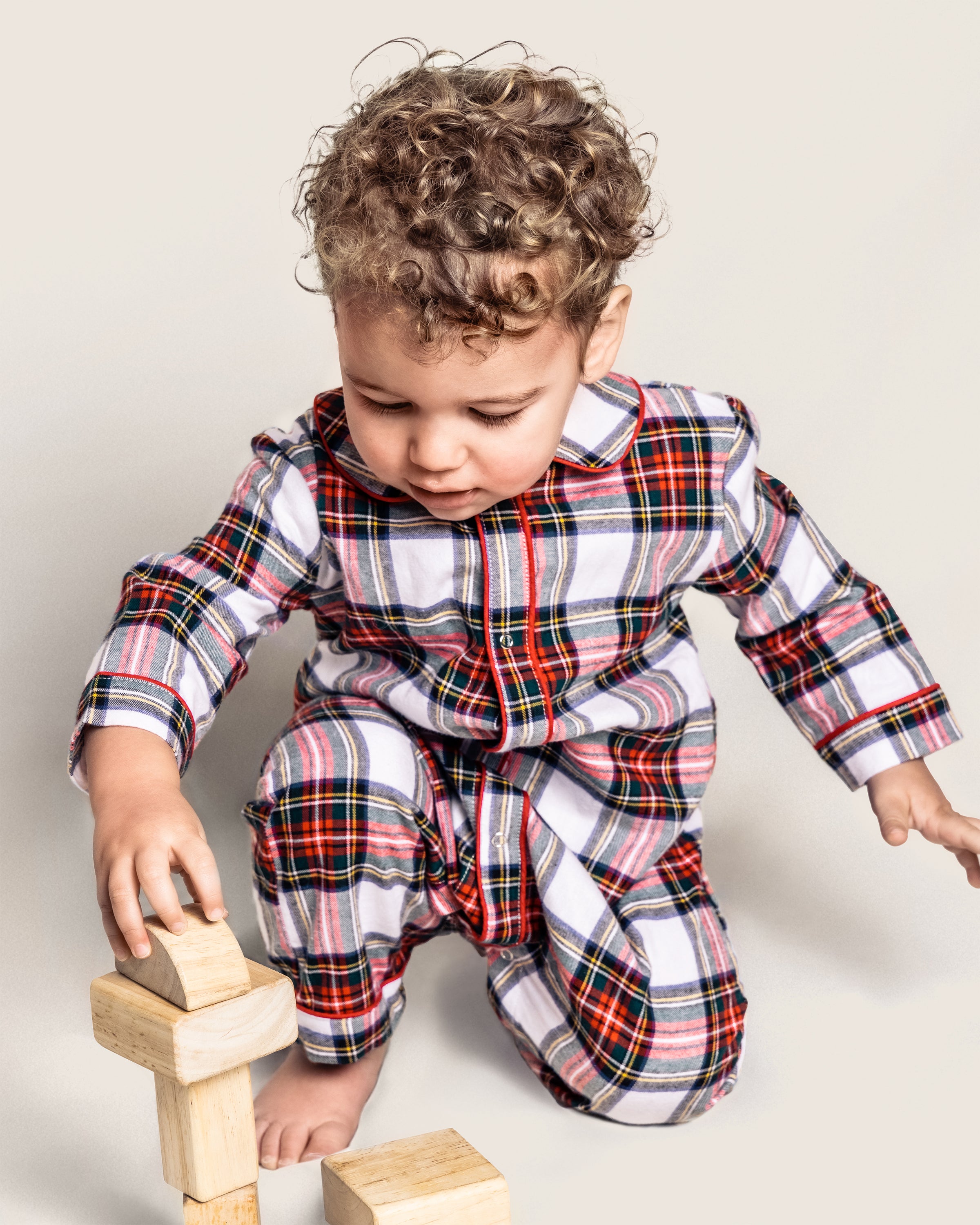A curly-haired toddler wears a Petite Plume Babys Brushed Cotton Cambridge Romper in Balmoral Tartan, reminiscent of Scottish tradition, while carefully balancing wooden blocks on the floor against a plain, neutral background.