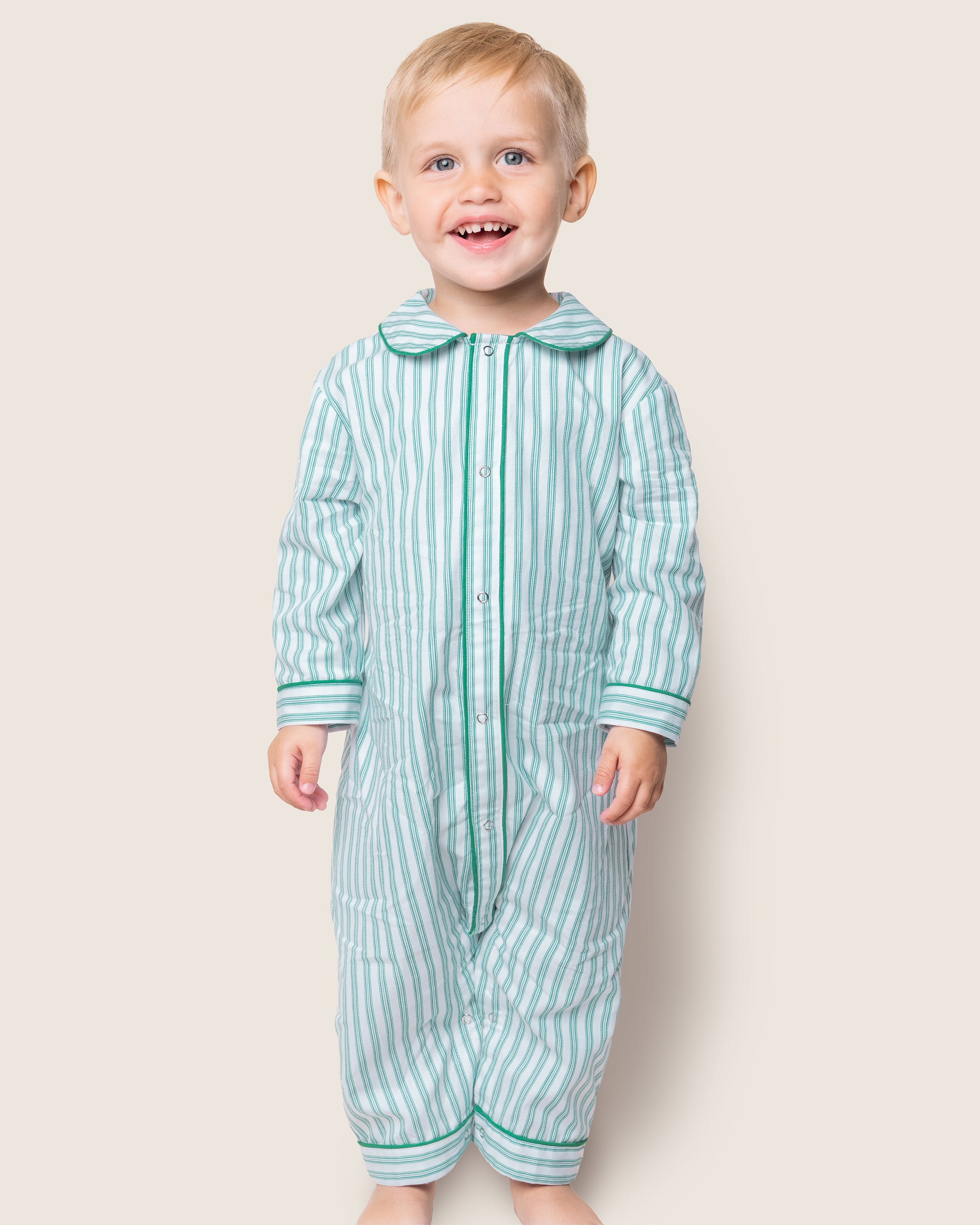 The smiling toddler is wearing Petite Plumes Babys Twill Romper in Emerald Ticking, showcasing a striped green and white design. This cozy sleepwear stands out against the plain light background, reminiscent of a classic French ticking pattern.