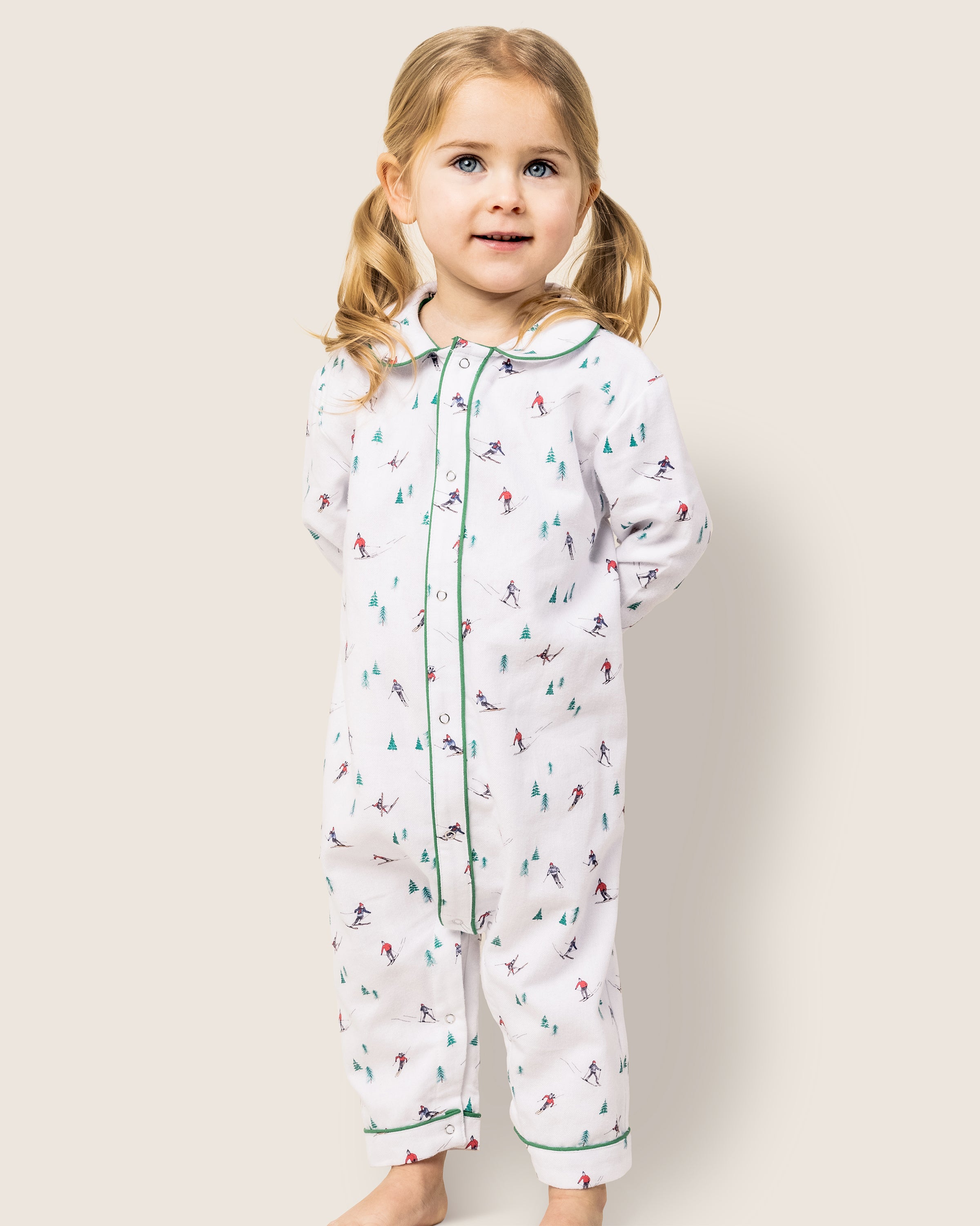 A child with blonde pigtails wears the Babys Flannel Cambridge Romper in Après Ski by Petite Plume, featuring small figures and trees with green piping. The child stands against a plain background, smiling brightly.