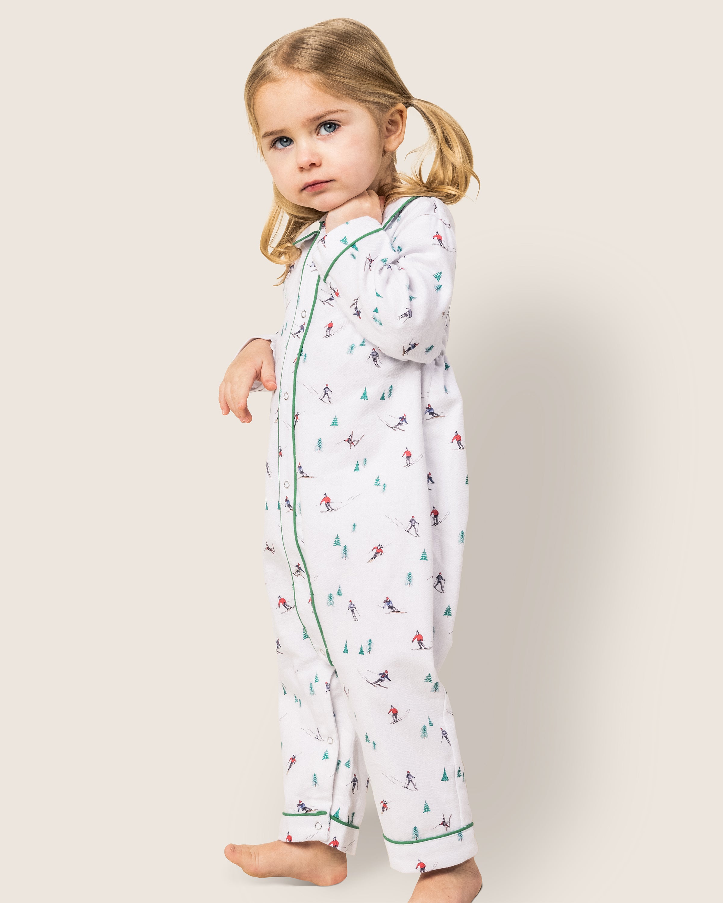 A young child with blonde pigtails wears a Petite Plume Babys Flannel Cambridge Romper in Après Ski, featuring a whimsical skier and tree pattern. The child stands barefoot against a plain background, gazing at the camera.
