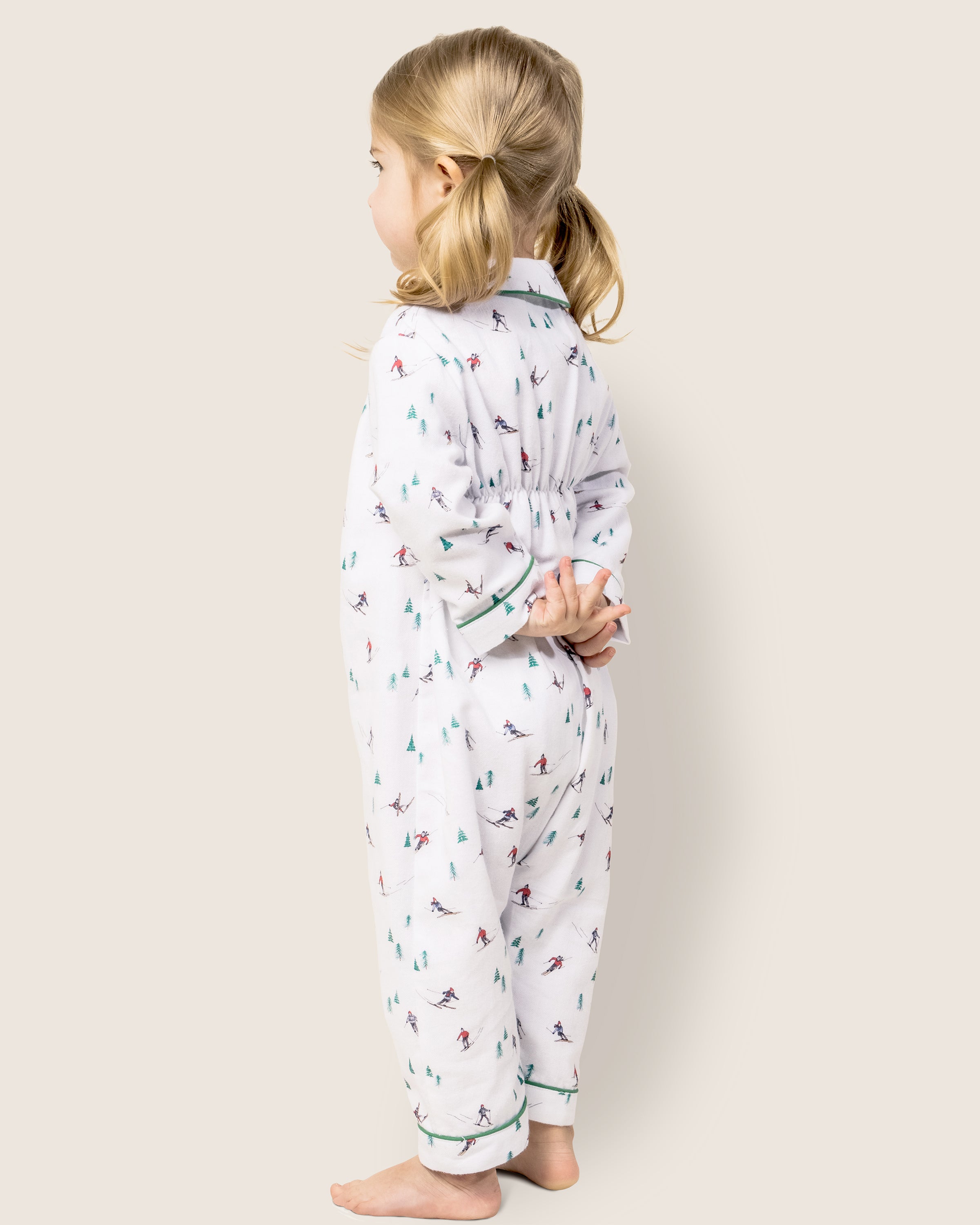A bare-footed child with blonde pigtails stands against a plain backdrop, wearing Petite Plumes Babys Flannel Cambridge Romper in Après Ski, hands clasped behind them as they look to the side.