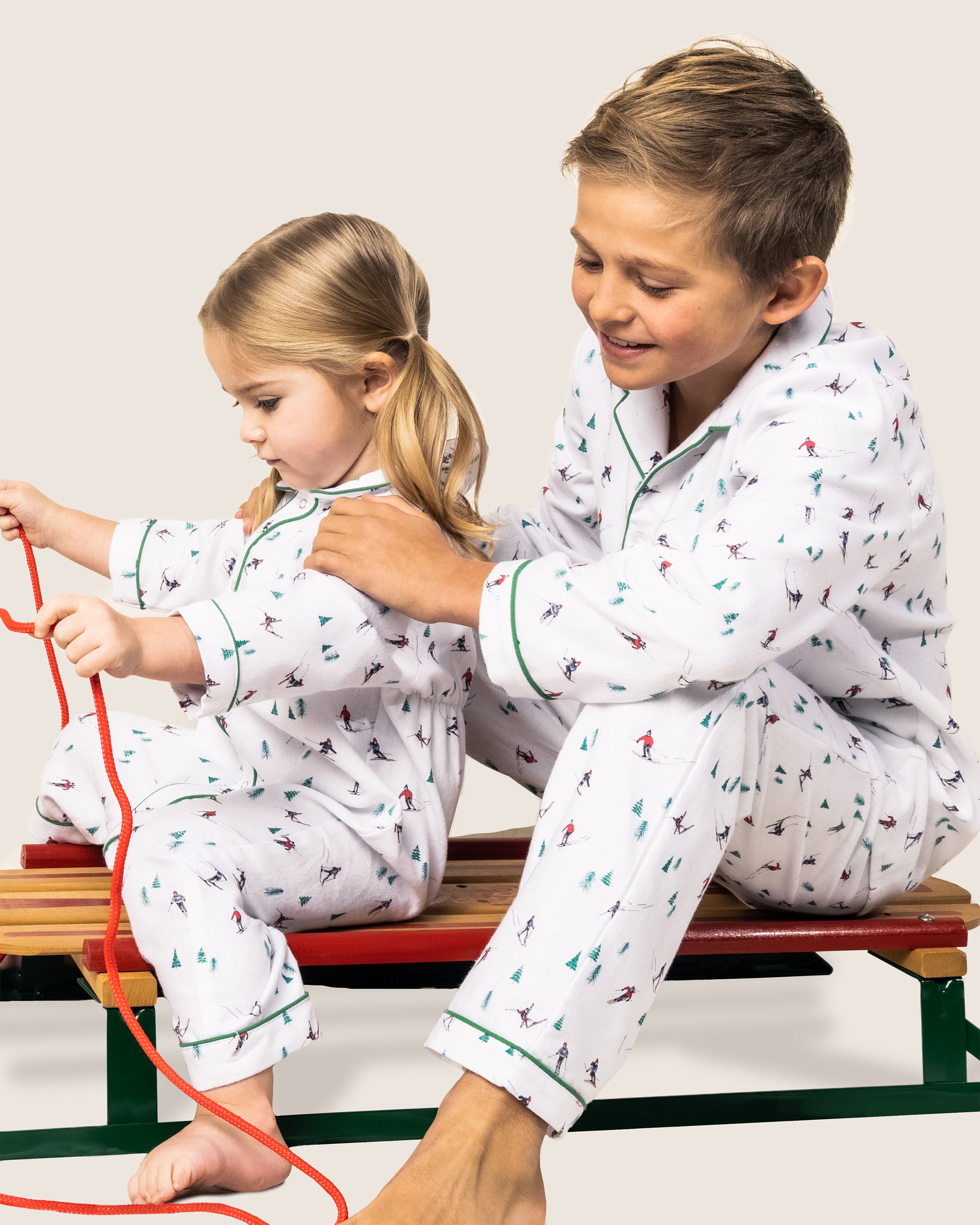 A young boy and girl in matching Babys Flannel Cambridge Rompers by Petite Plume with Après Ski prints sit on a wooden sled. The boy smiles while the girl holds a red rope, looking content and playful.