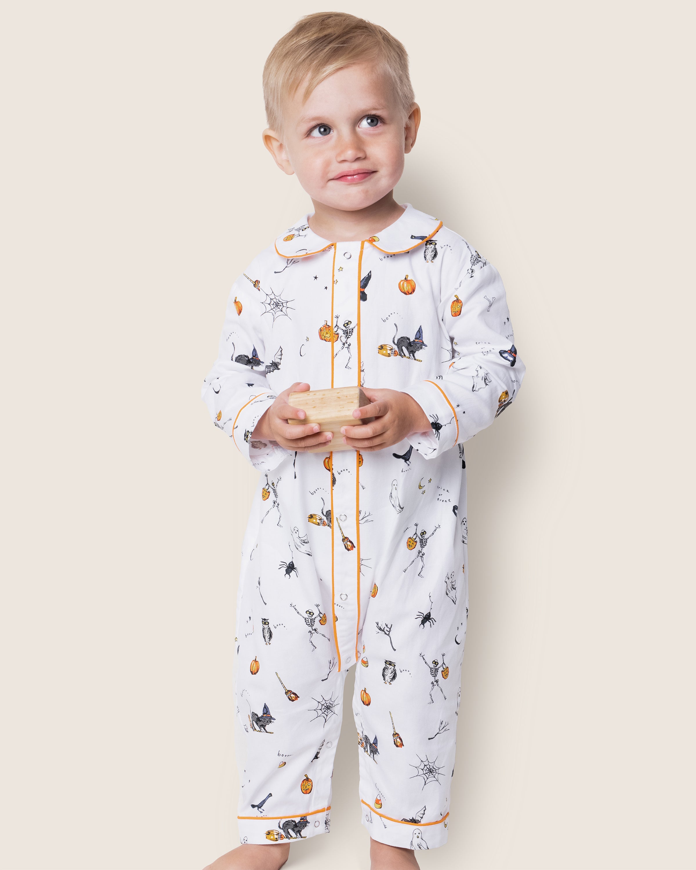 A child wearing Petite Plumes Babys Twill Cambridge Romper in Trick or Treat, decorated with pumpkins, witches, and cats, holds a small wooden block. The flame-retardant onesie with orange trim is set against a plain light background as the child gazes upward with a slight smile.