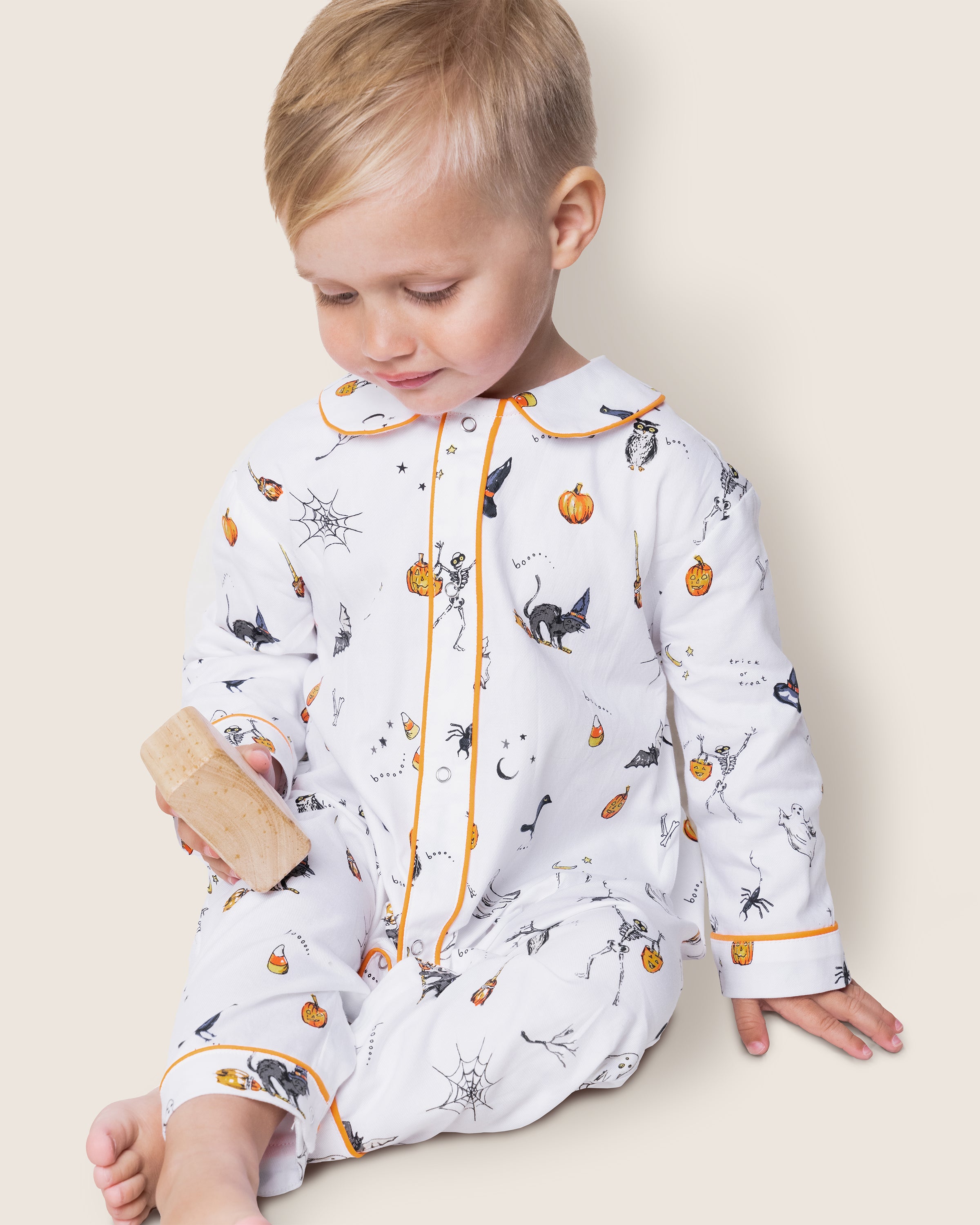 A toddler in Petite Plumes Babys Twill Cambridge Romper, Trick or Treat, sits adorned with Halloween icons like cats, pumpkins, and webs, holding a wooden block and capturing the festive spirit.
