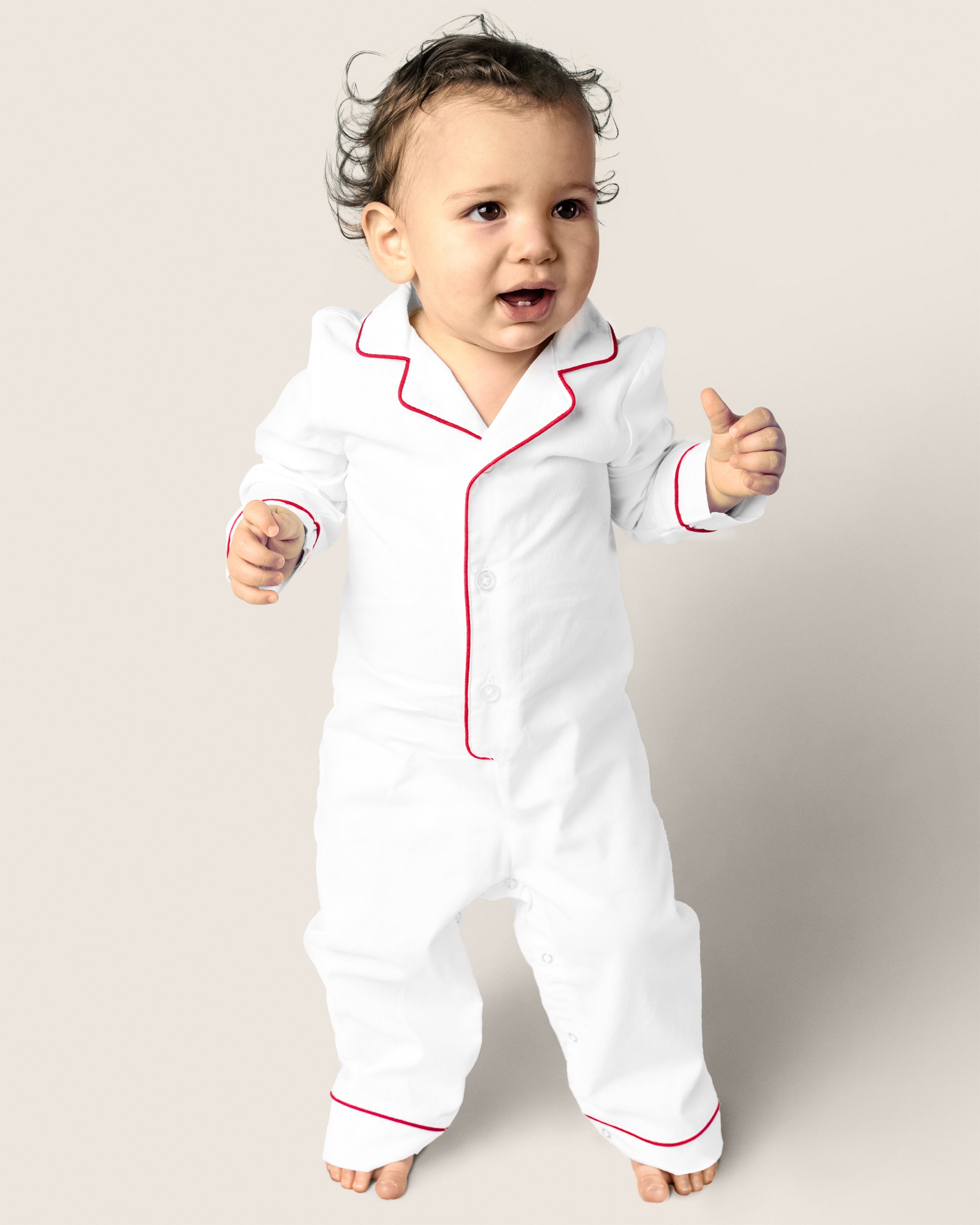 A toddler in Petite Plumes Babys Twill Romper in White with Red Piping stands against a plain background, looking up curiously with slightly tousled hair, as if just woken from sleepwear comfort.