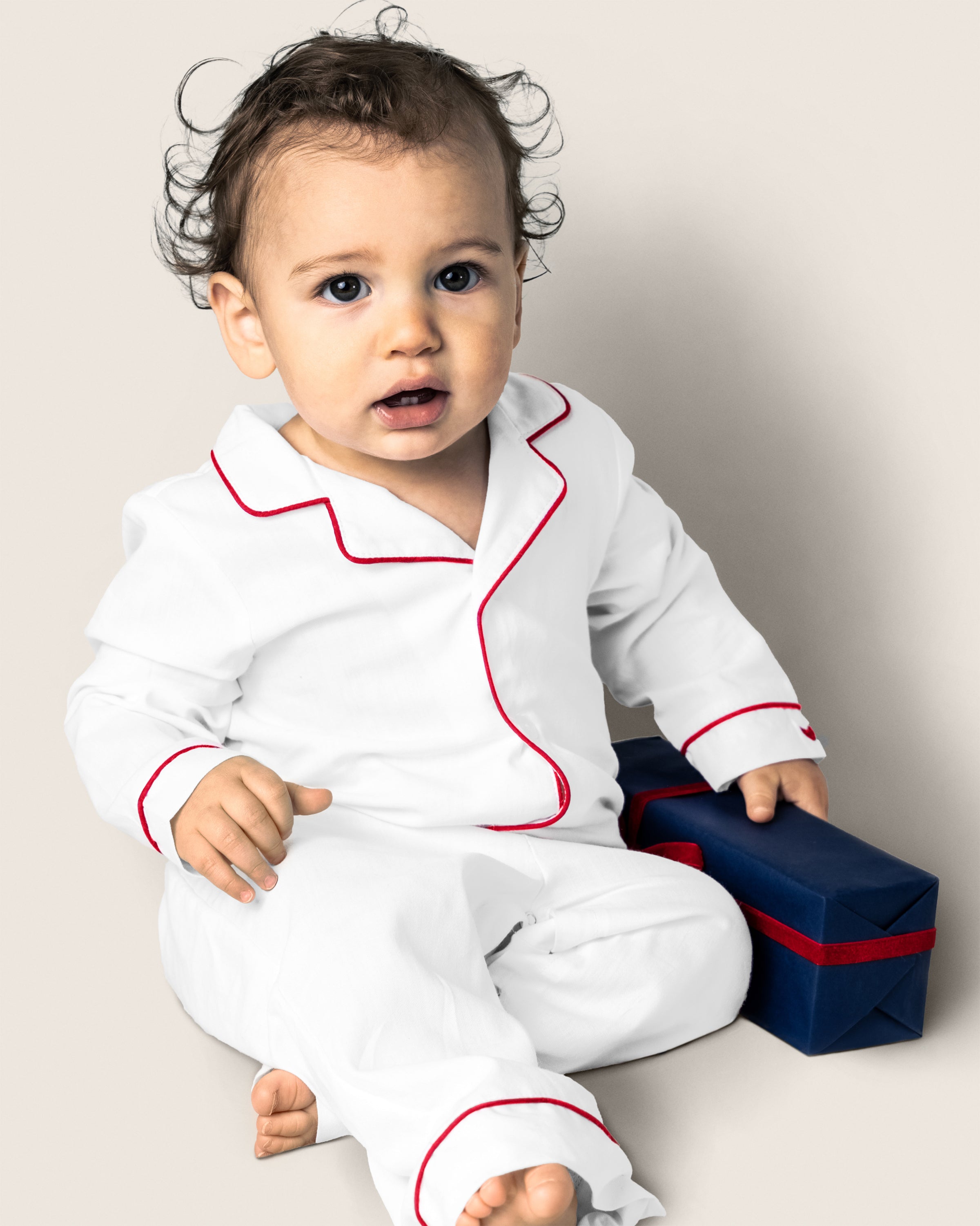 A baby in a Petite Plume Babys Twill Romper in White with Red Piping sits barefoot, holding a finger to the side. A blue gift box with a red ribbon adds charm against the neutral-toned background. This cozy sleepwear is crafted from brushed fabric for ultimate comfort.