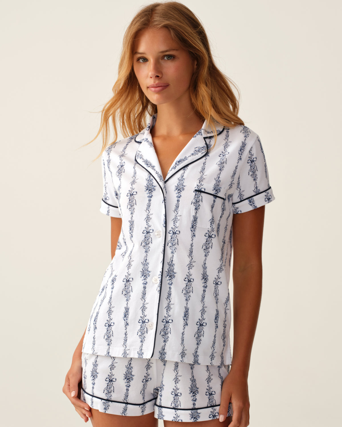 A woman wears the Greek Embroidered Poppy Pima Short Set in Navy Mayfair Toile—featuring a white pajama set with blue floral vertical stripes, including a short-sleeve button-up top and matching shorts—as she stands against a neutral background.