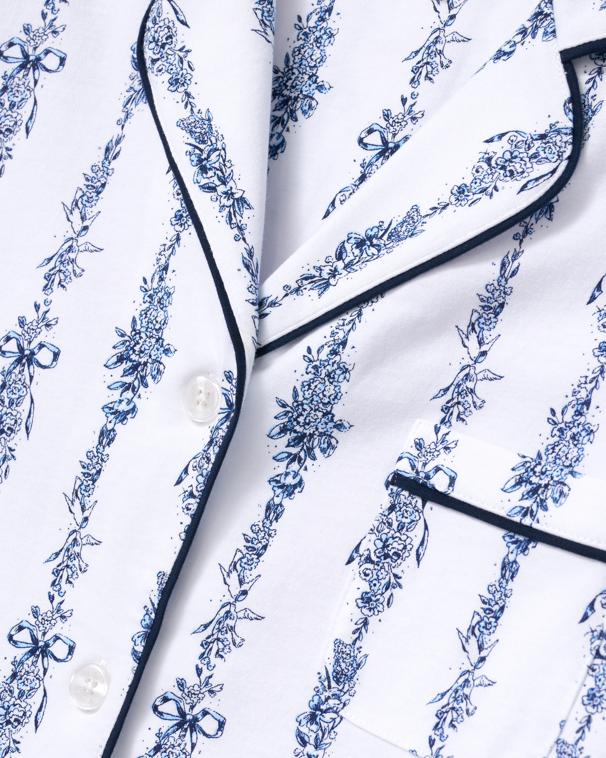 Close-up of the Greek Embroidered Poppy Pima Short Set in Navy Mayfair Toile, featuring a white pajama top with blue floral and bow patterns, white buttons, black piping, and a chest pocket.