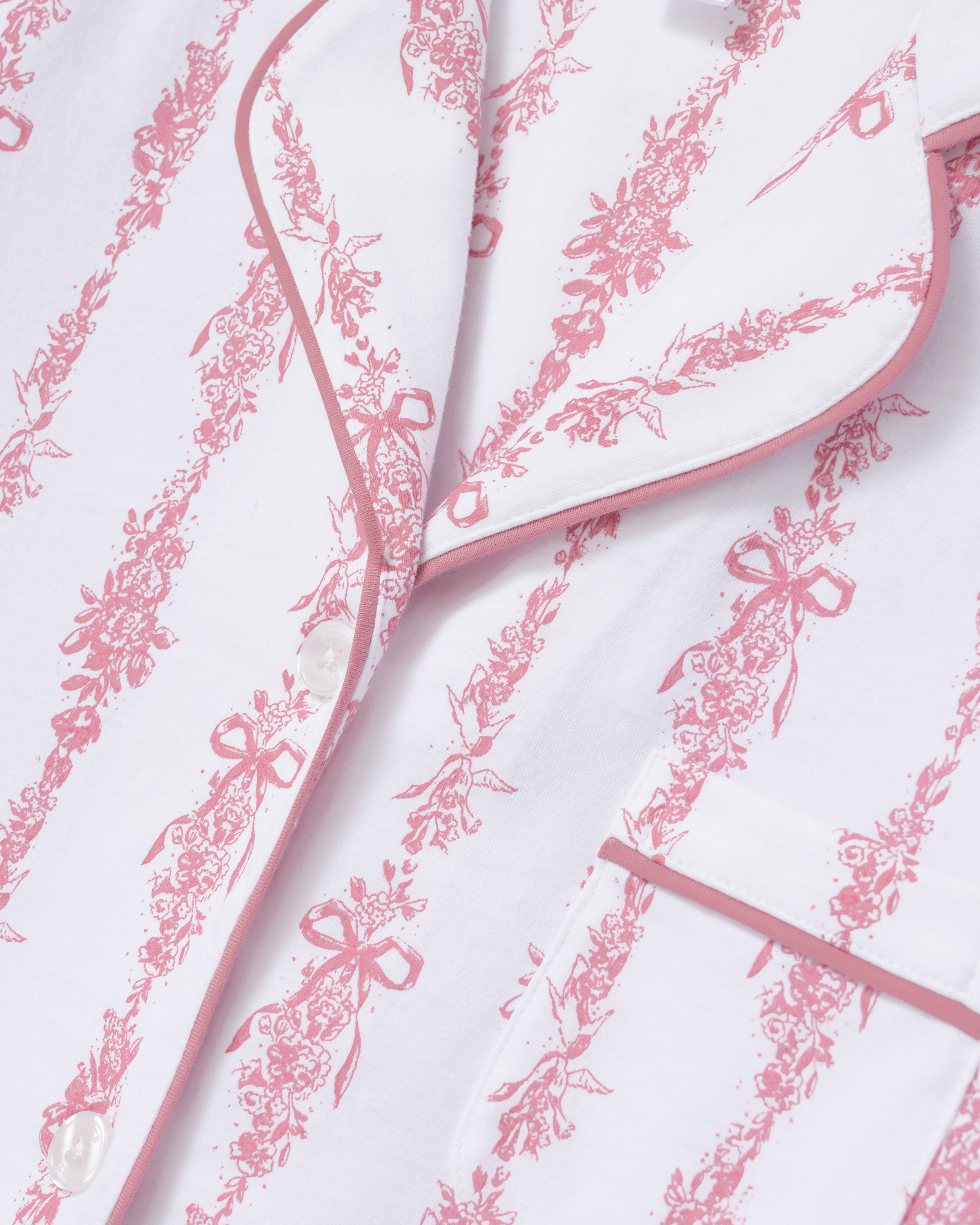 Close-up of The Poppy Pima Short Set in Pink Mayfair Toile, featuring a white pajama shirt adorned with pink floral and bow patterns, pink piping along the collar and pocket, and finished with white buttons.