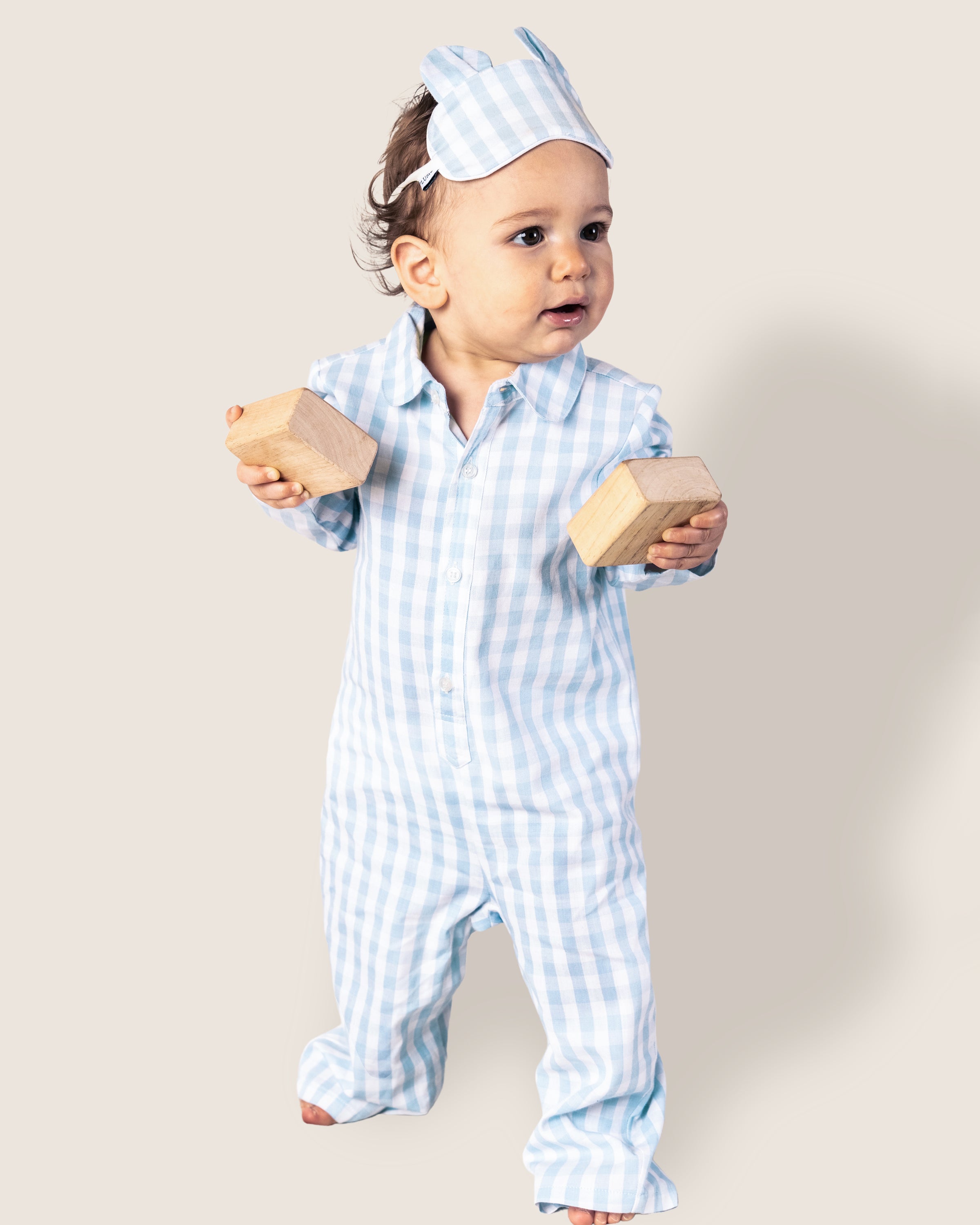 A baby in a Petite Plume Babys Twill Romper in light blue gingham with a matching eye mask holds two wooden blocks while standing against a plain, light-colored background.