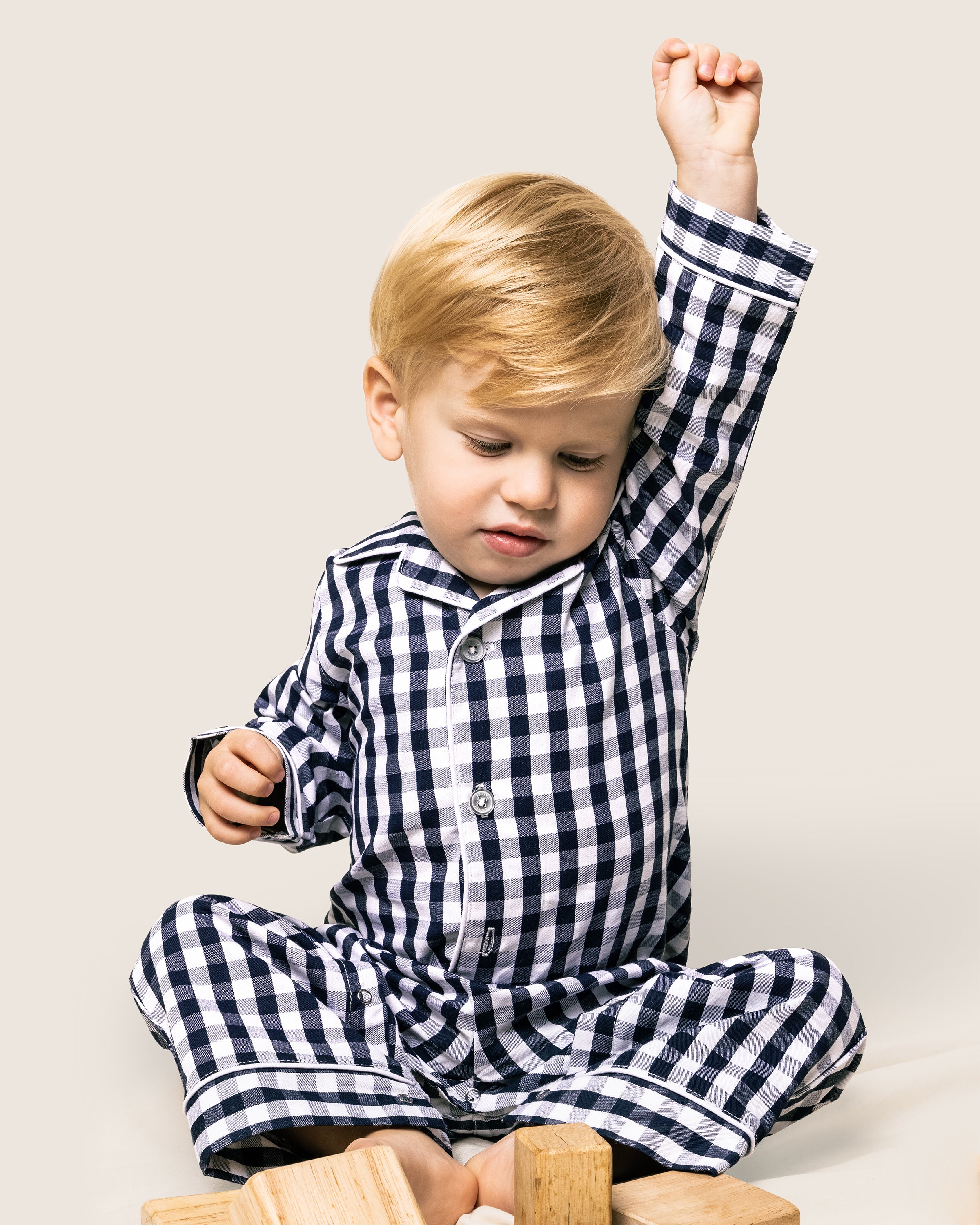 A blonde toddler sits wearing Petite Plumes Babys Twill Winter Romper in Navy Gingham. One arm is raised, and the child looks down with a slight smile. Wooden blocks are scattered in front of them.