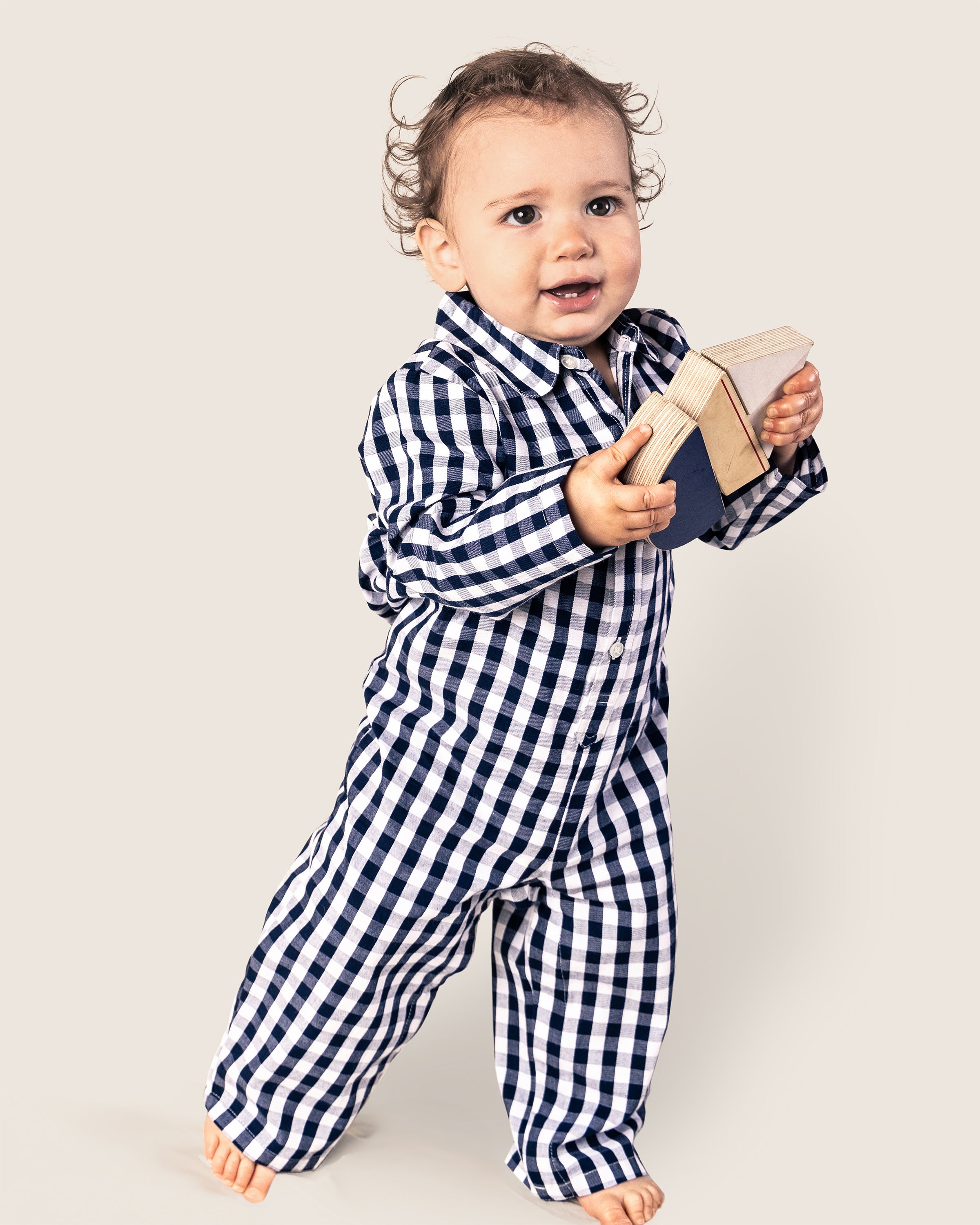 A toddler in a Petite Plume Babys Twill Winter Romper in Navy Gingham plays with wooden blocks, walking barefoot against a plain, light-colored background.