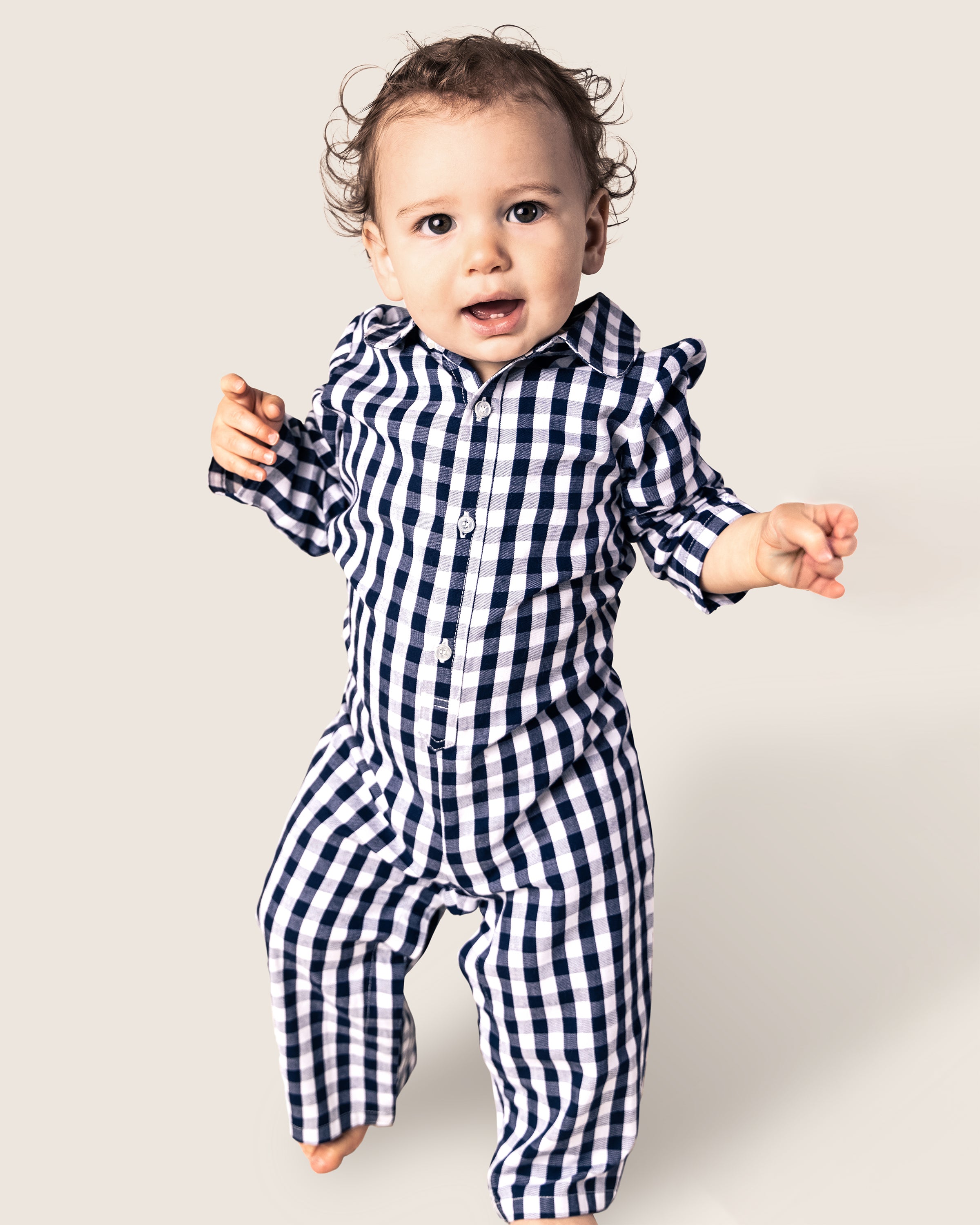 A toddler in a navy gingham Babys Twill Winter Romper by Petite Plume stands with arms slightly raised, looking at the camera with a neutral expression against a plain beige background.