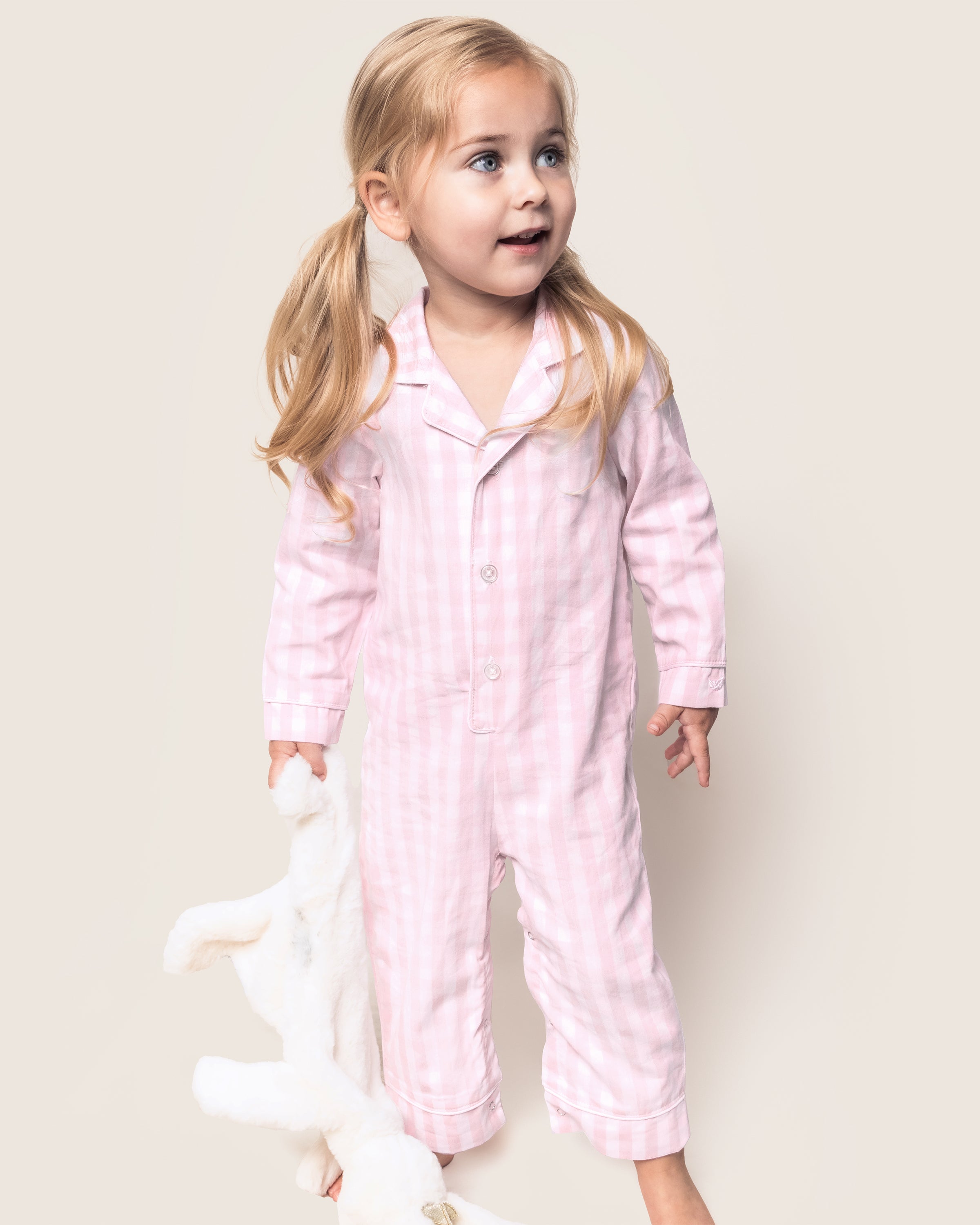 A young blonde child with pigtails wears a Petite Plume Babys Twill Romper in Pink Gingham while holding a white stuffed animal against a light-colored background.