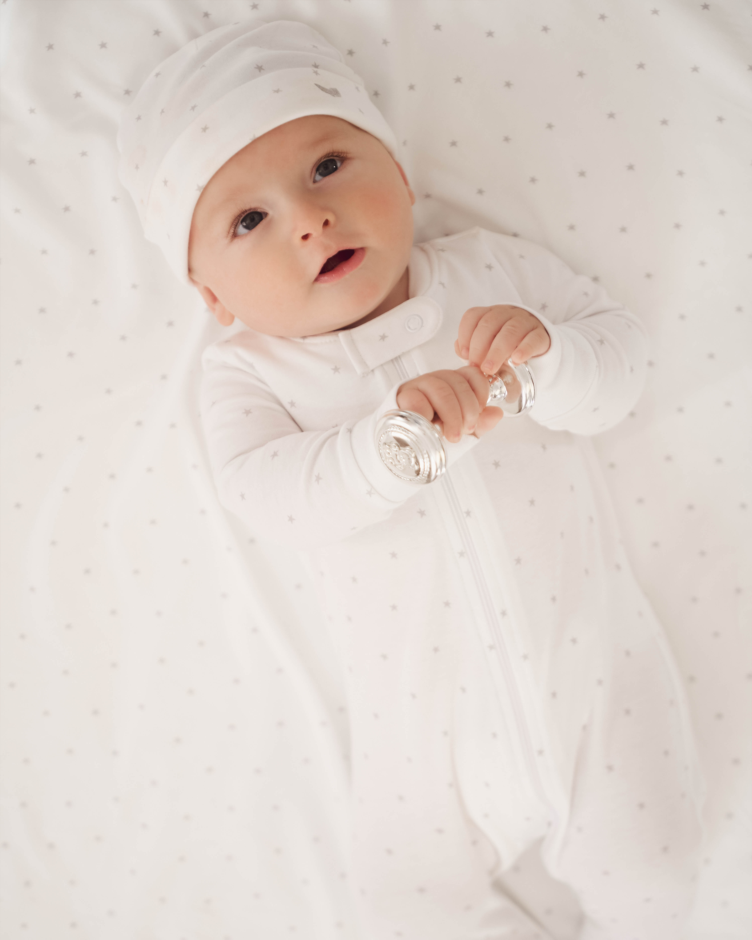 A baby in the Welcome Home Baby Set - Grey Stars by Petite Plume, featuring a white romper and hat with delicate polka dots, lies on a matching blanket holding a silver rattle. The baby gazes upward, creating a soft, peaceful ambiance.