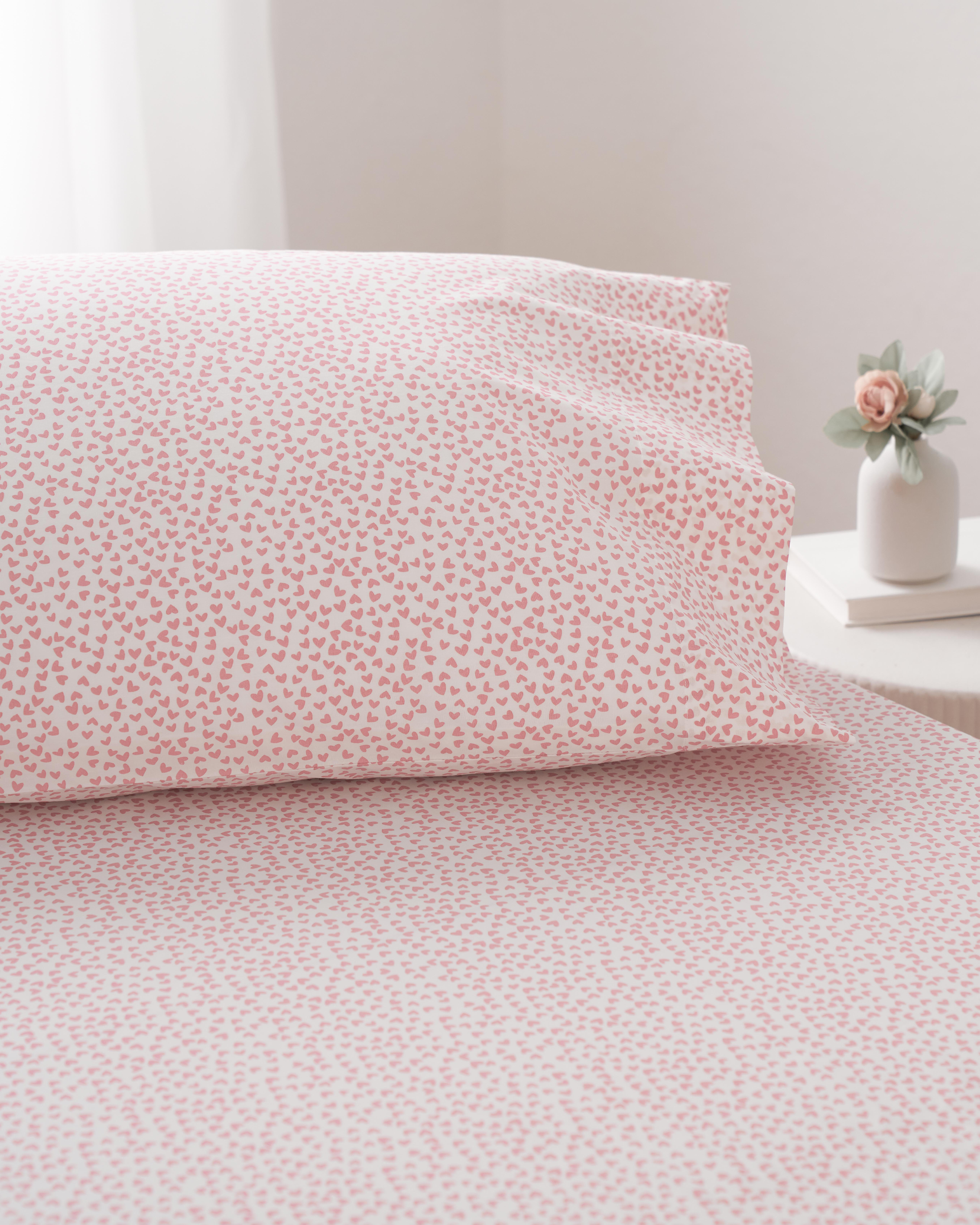 A bed is adorned with Petite Plumes Luxe Premium Cotton Sweethearts Bed Sheets, featuring pink and white hearts, and a matching pillowcase. A small vase with a pink flower sits on the bedside table. Natural light illuminates through the window with white curtains, creating a cozy setting.