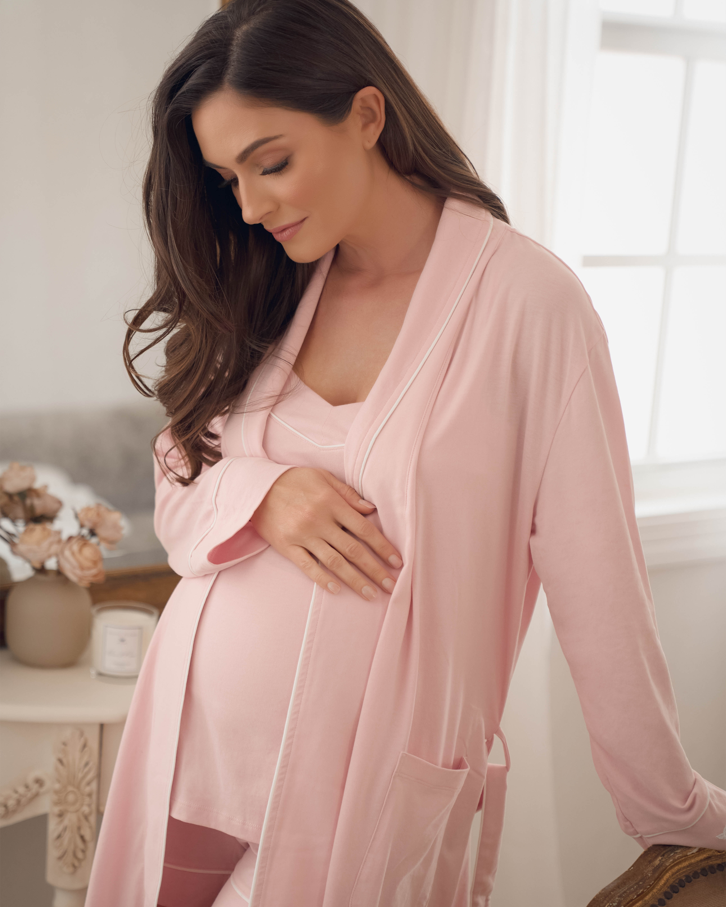 A pregnant woman with long brown hair stands by a window, gently touching her belly while wearing a Pink Womens Pima Maternity Robe by Petite Plume. The softly lit room, adorned with flowers and a cream-colored table, exudes serenity and warmth.
