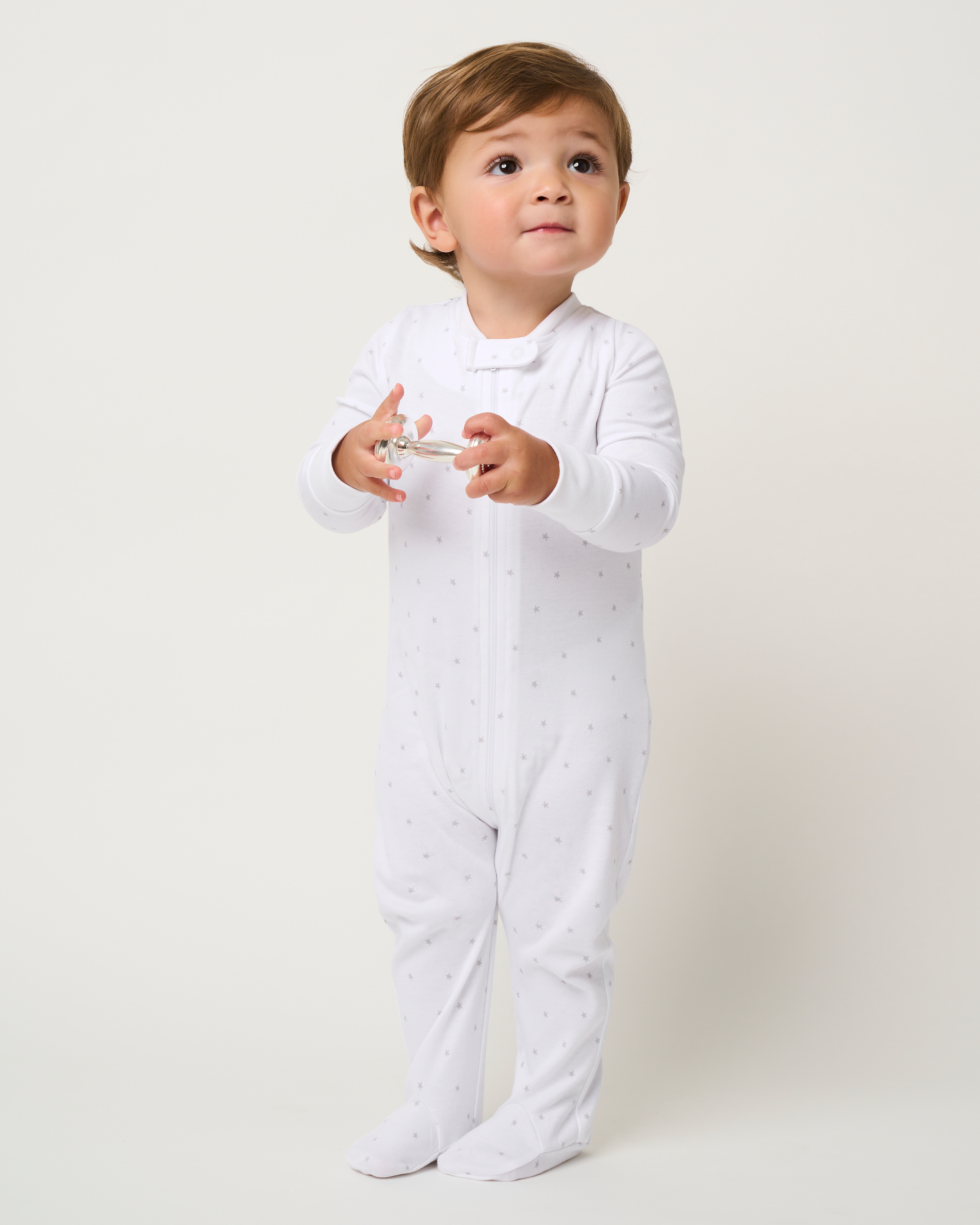 A toddler in Petite Plumes Babys Pima Romper in Grey Stars, a white full-body romper with gray dots, stands against a plain background holding a clear pacifier and gazing slightly upward. Made from soft Pima cotton, this sleepwear combines comfort and style.