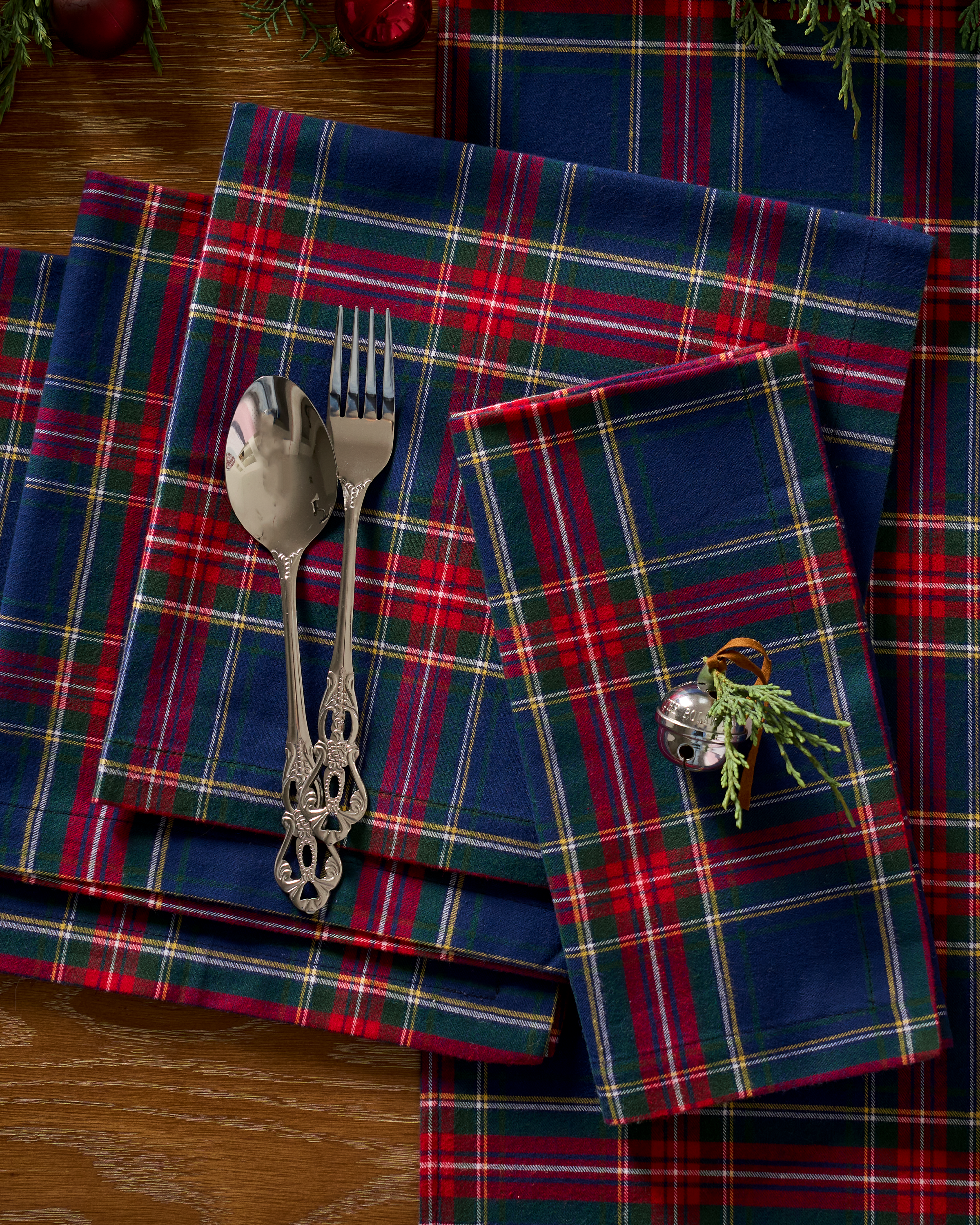 Petite Plumes Signature Twill Napkins in Windsor Tartan feature a festive red, green, and blue plaid made from 100% cotton, topped with silver cutlery, including a spoon and fork. A silver bell decoration with greenery enhances the folded napkins on the wooden surface for a holiday ambiance.