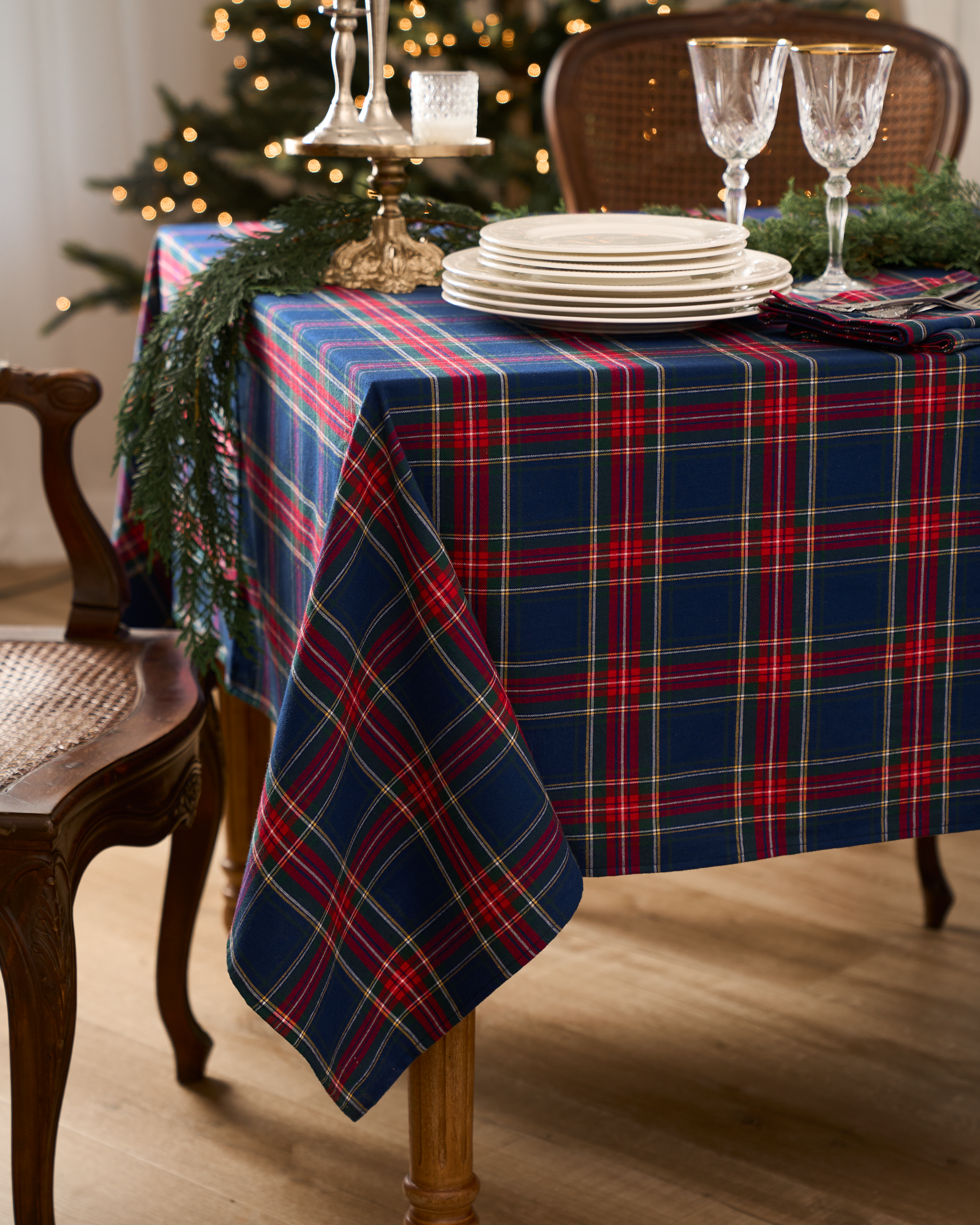 A dining table flaunts a festive tablescape with the Petite Plume Signature Twill Tablecloth in Windsor Tartan, complete with white plates, crystal glasses, and a gold candlestick. Green pine branches add charm while a Christmas tree glows warmly in the background.