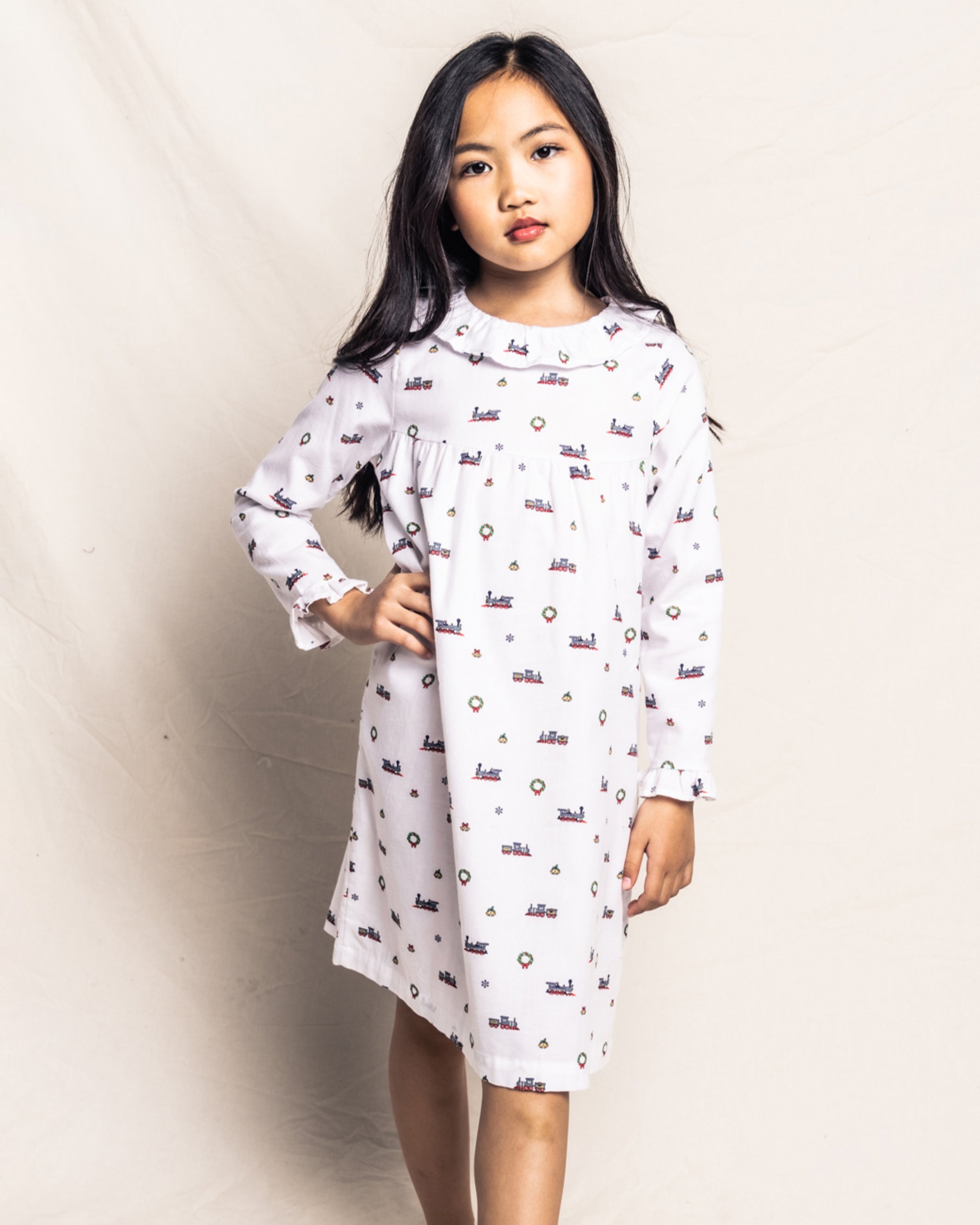 A young girl with long dark hair poses confidently in Petite Plumes Girls Twill Scarlett Nightgown in Arctic Express, featuring a white, long-sleeve design adorned with colorful patterns. The cozy-soft twill fabric enhances her comfort and elegance.