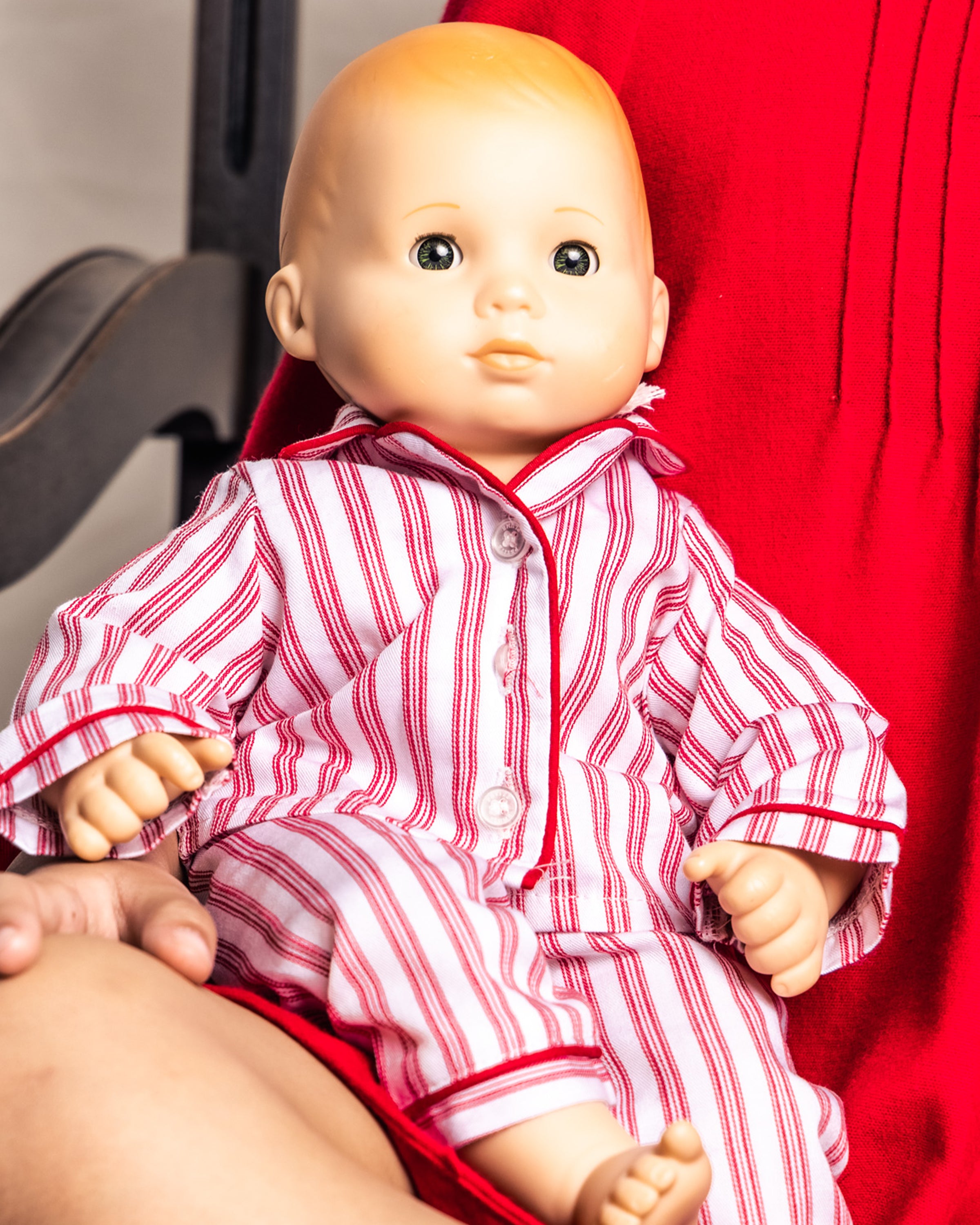 A bald 18 doll in Petite Plumes Doll Pajamas in Antique Red Ticking sits on a lap, both donning matching red attire for a delightful look that lets them align perfectly with their cherished doll.