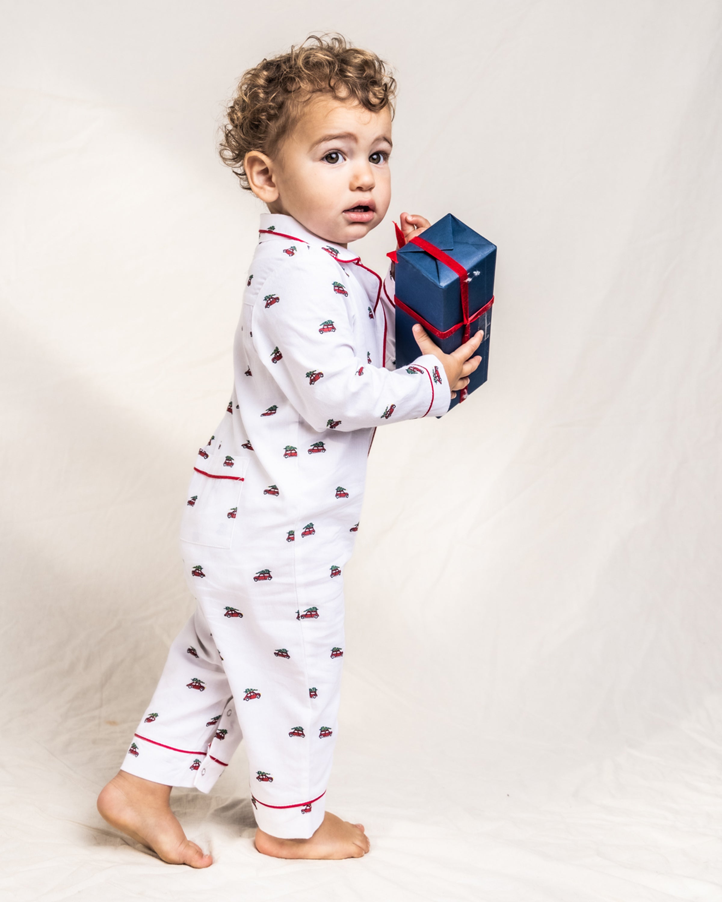 A curly-haired toddler stands barefoot in a luxurious Babys Twill Romper in Holiday Journey by Petite Plume, holding a blue gift box with a red ribbon and looking at the camera against a plain background.