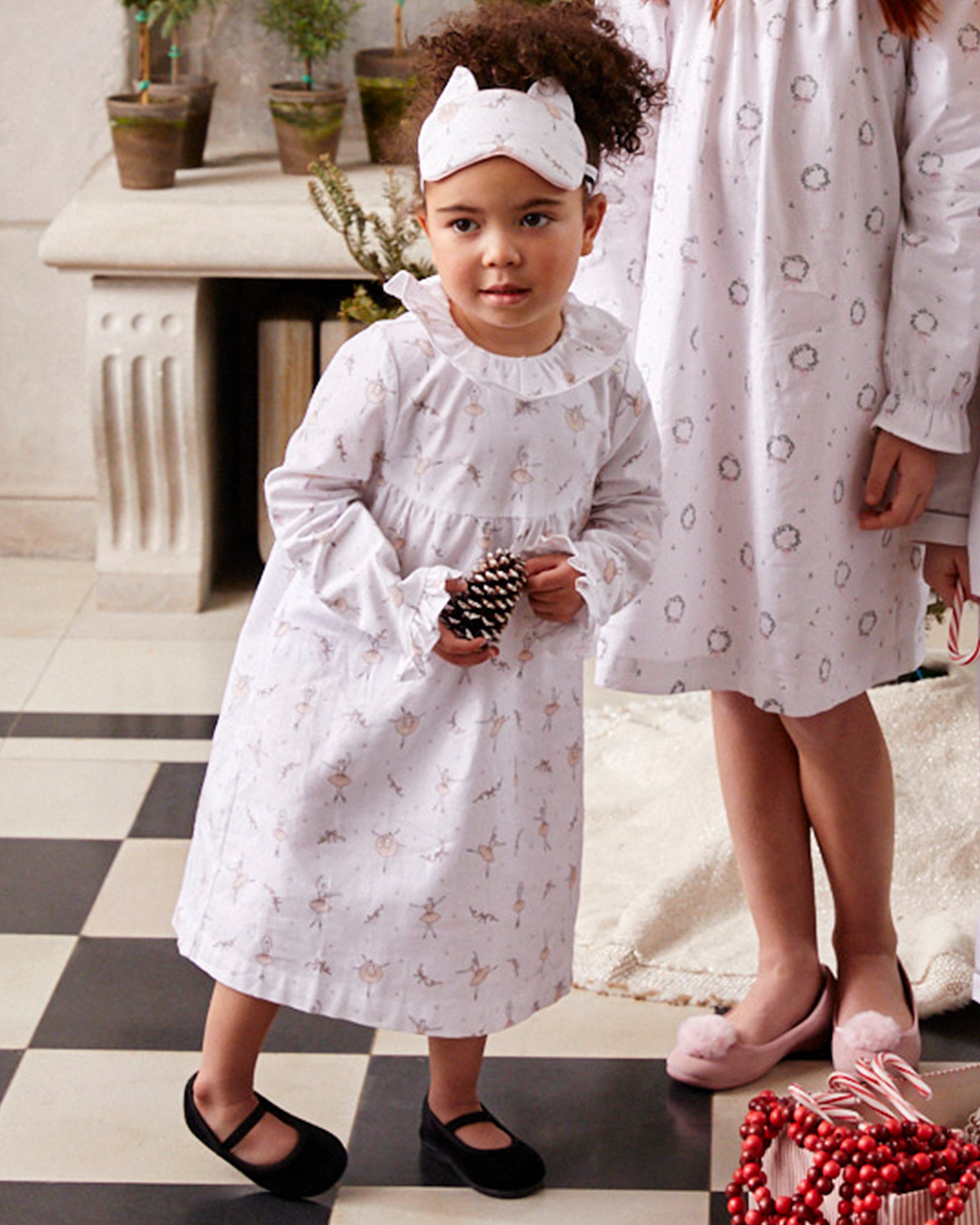 A little girl in a white printed nightgown and matching headband stands holding a toy, wearing Petite Plumes Kids Delphine Slippers in black velvet. Next to her, an older child in a similar gown is partially visible. The room features a checkered floor and a white fireplace.