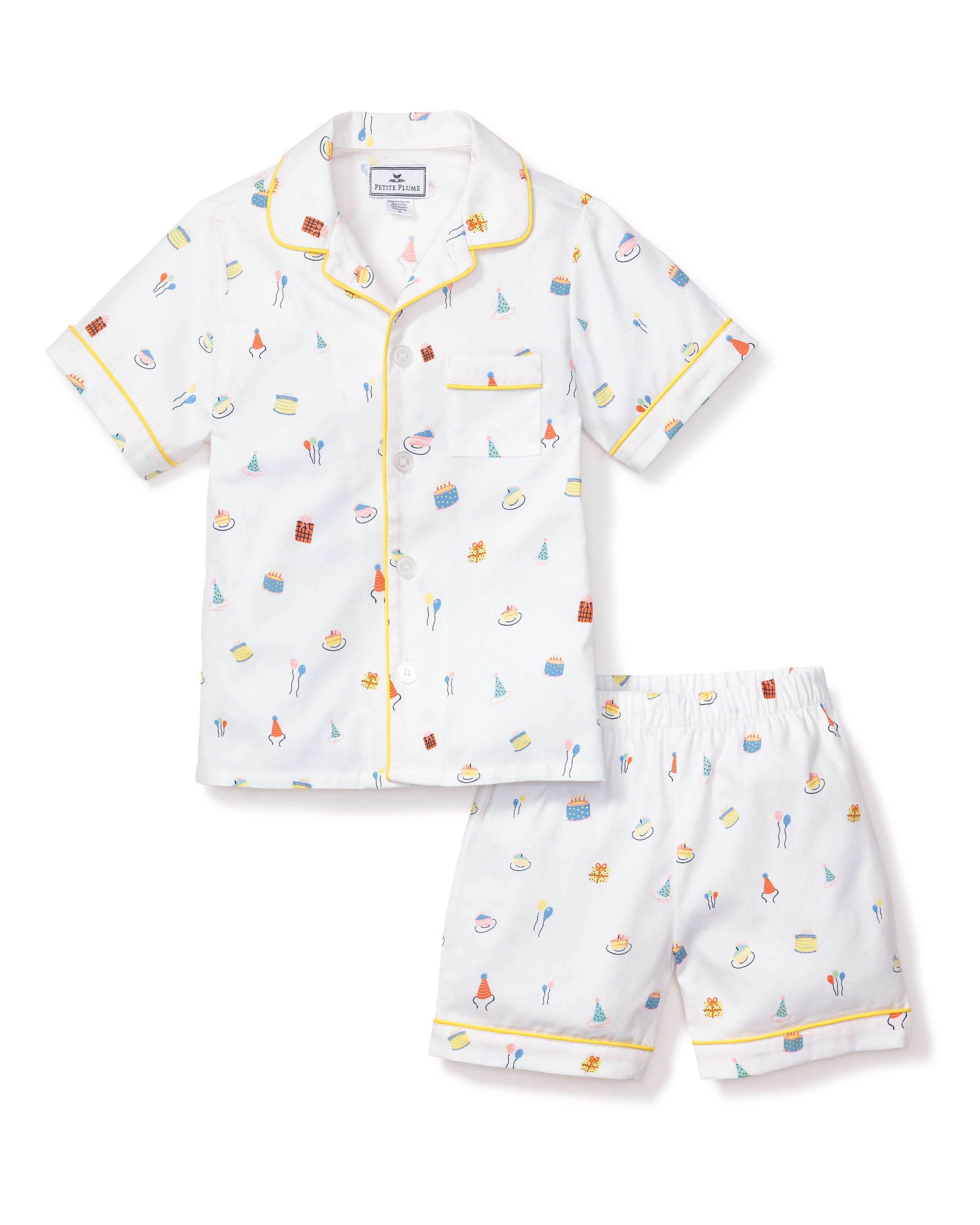 The Petite Plume Kids Twill Pajama Short Set in Birthday Wishes includes a short-sleeve, button-up shirt and shorts. The white fabric is decorated with colorful birthday fantasy prints of cakes, balloons, and party hats, featuring luxurious yellow piping on the cuffs and collar.