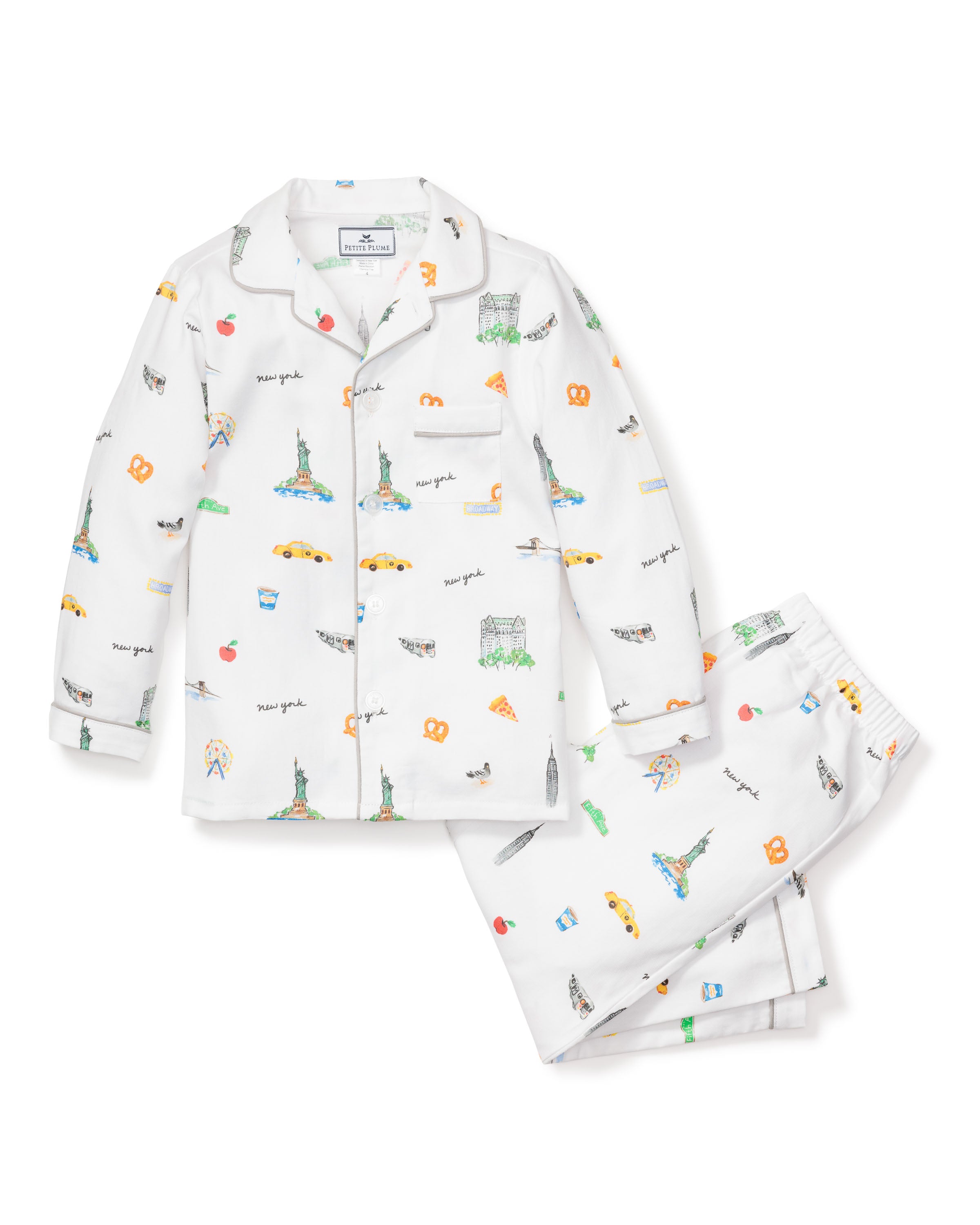 The Kids Twill Pajama Set in New York! New York! by Petite Plume features white pajamas with colorful prints of the Statue of Liberty, Empire State Building, taxis, and Brooklyn Bridge. The set includes a button-up shirt and pants adorned with pizza slices, pretzels, and New York script.