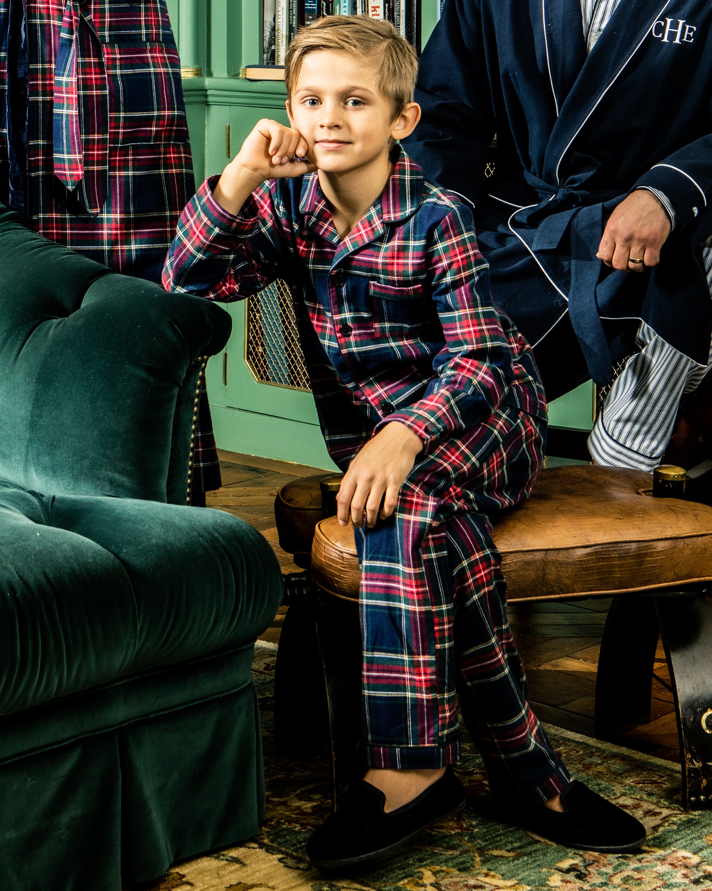 A young boy wears plaid pajamas and sits on a stool, resting his chin on his hand with a relaxed expression. Behind him, a figure in a robe stands near a green couch and patterned rug. He complements his attire with Petite Plumes Kids Hamptons Loafer in black velvet for added style.