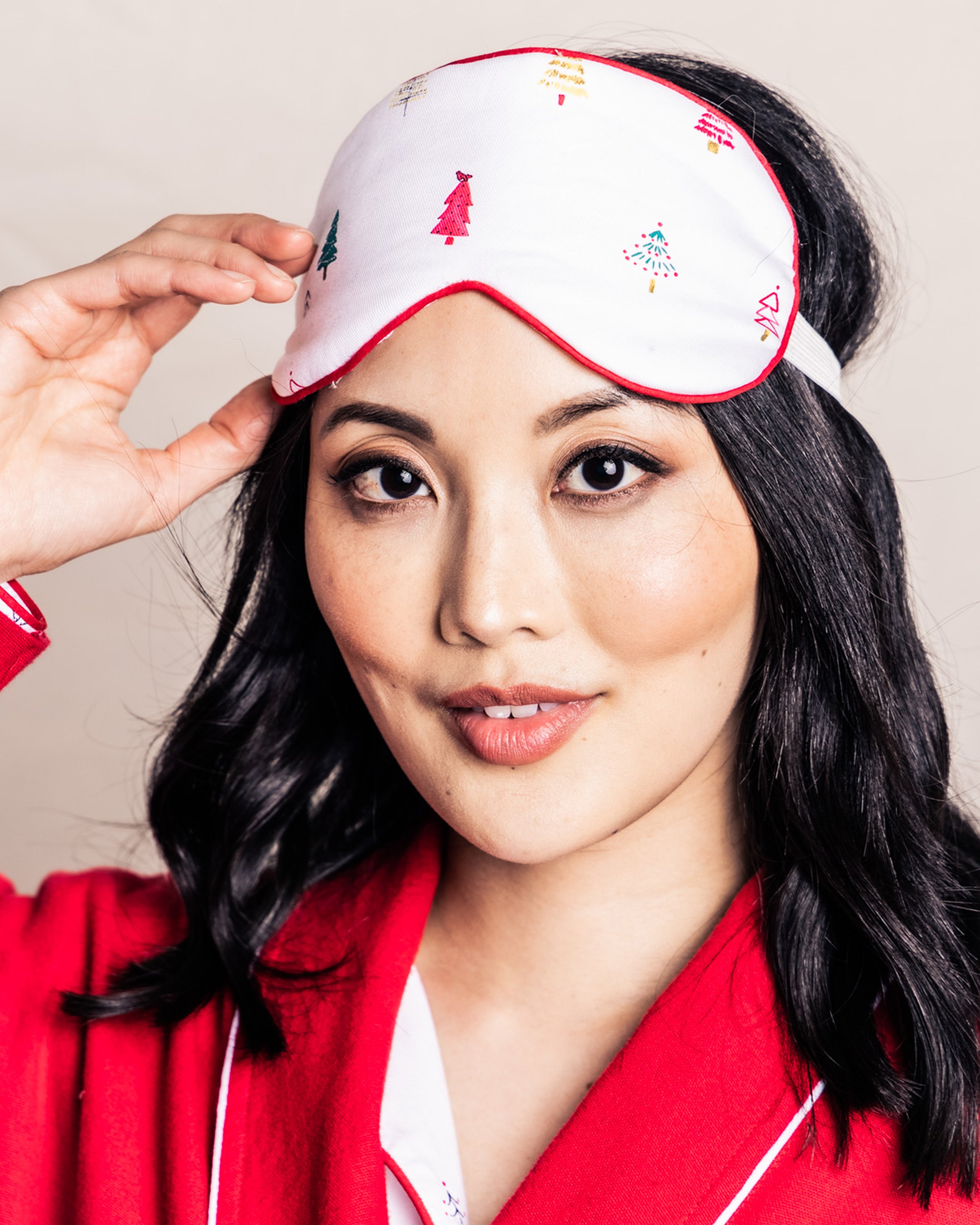 A woman with long black hair smiles while wearing a red pajama top and the Merry Trees sleep mask by Petite Plume. She gently touches the mask, showcasing it as a stylish sleepover accessory against a plain background.