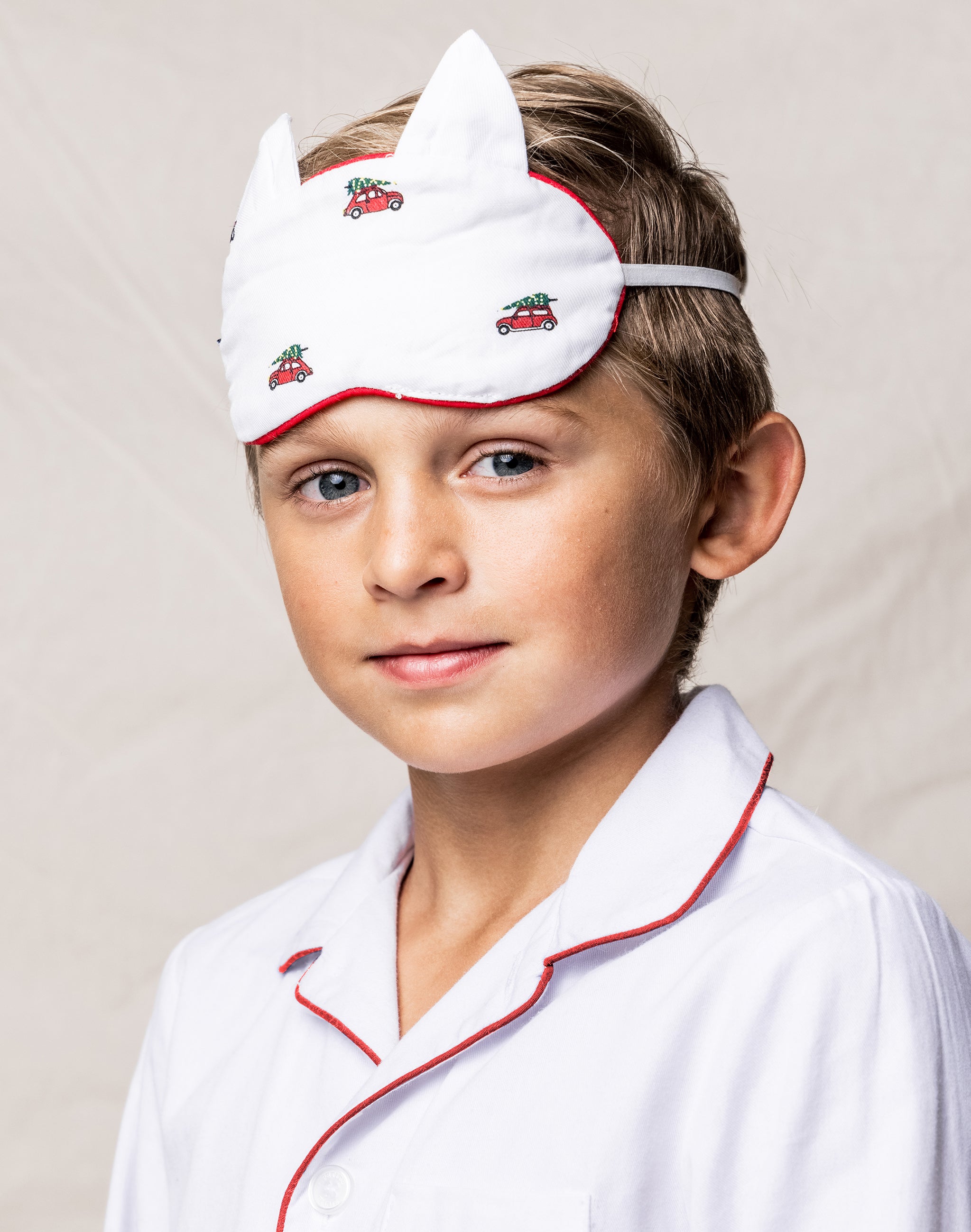 A young boy, in luxurious sleepwear from Petite Plume, wears the Kids Twill Kitty Sleep Mask in Holiday Journey with red car and tree designs. The mask perfectly matches his pajamas with red piping, adding charm against a soft light beige background.