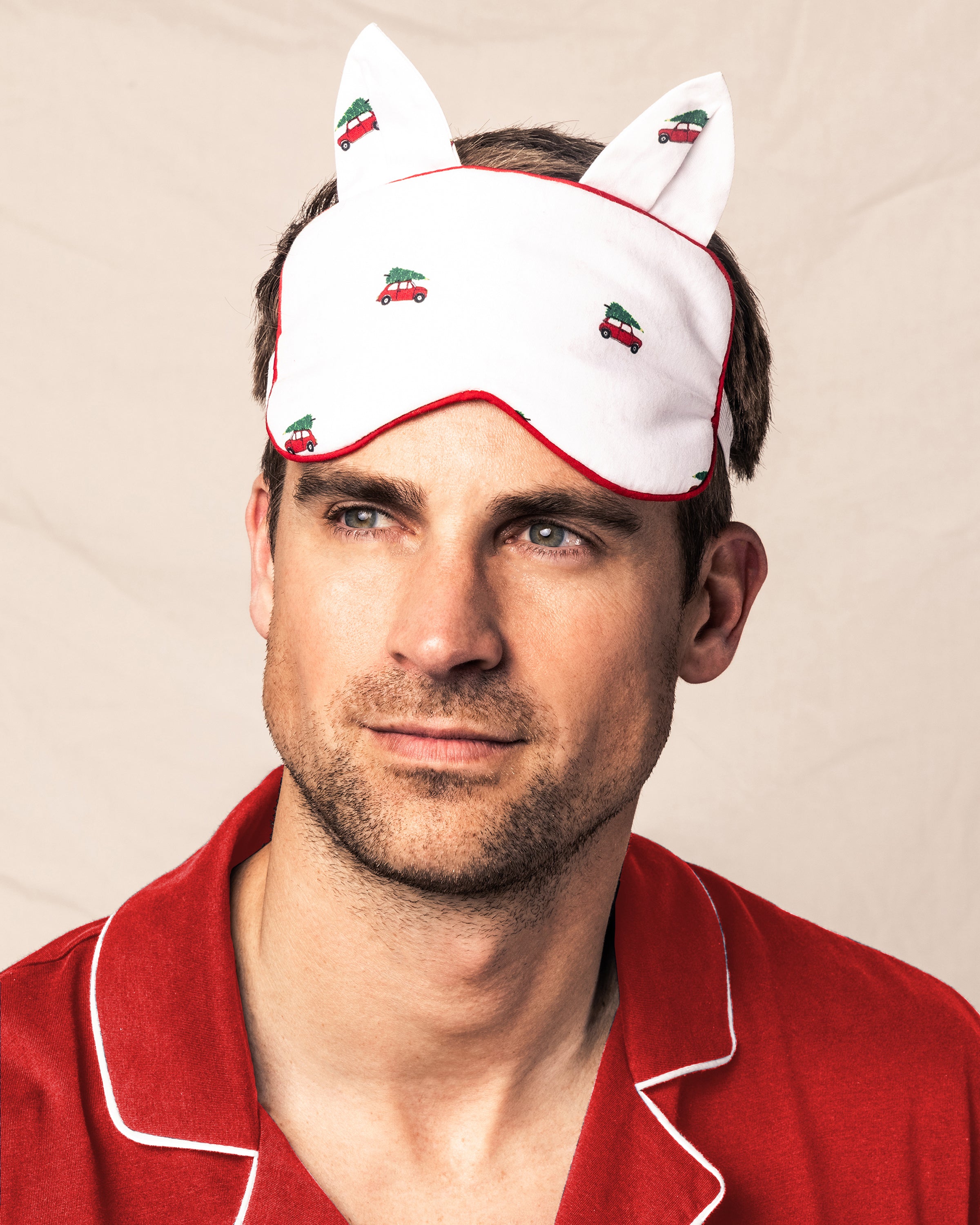 A man wearing red pajamas with white piping and a monogrammed Petite Plume Adults Twill Kitty Sleep Mask in Holiday Journey, featuring ears and small car designs, gazes to the side against a plain background.