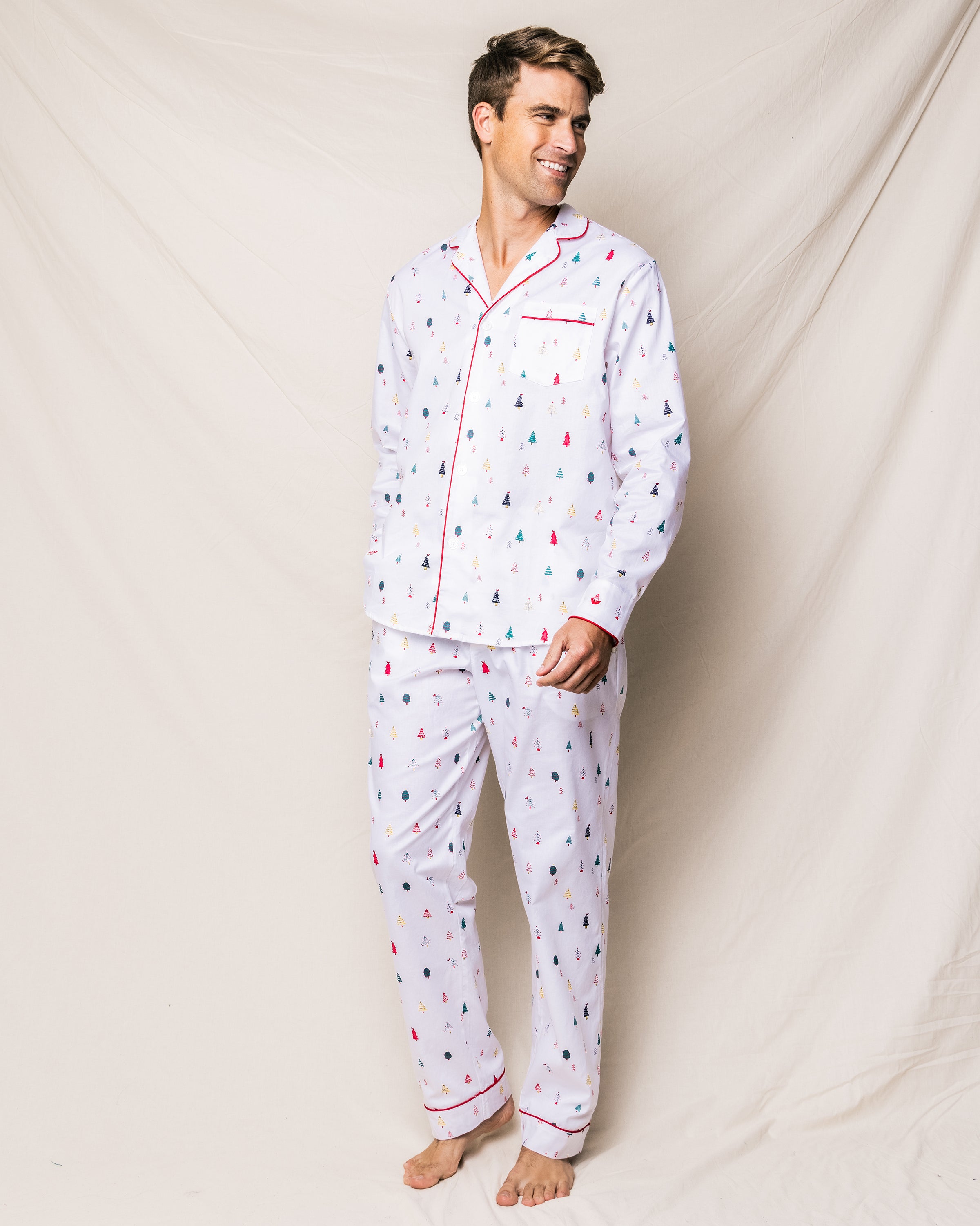 A man stands barefoot against a light beige fabric backdrop, wearing Petite Plumes Mens Twill Pajama Set in Merry Trees. The pajamas, featuring colorful tree patterns and red piping, showcase the luxurious sleepwear of finest quality cotton as he smiles joyfully to the side.