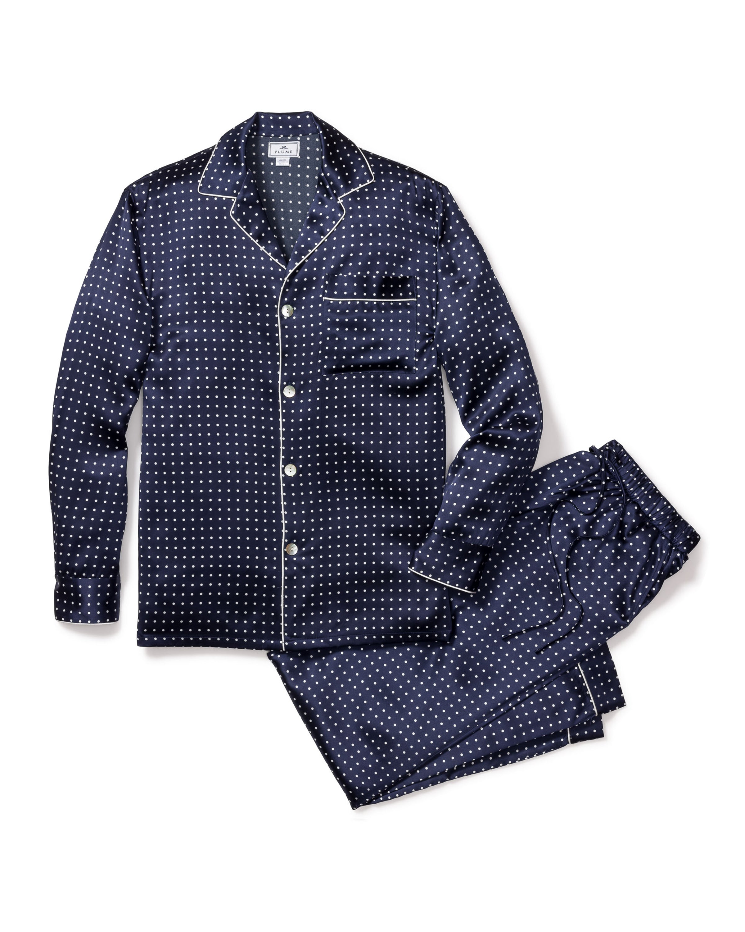 The Mens Silk Pajama Set in Navy Polka Dot by Petite Plume is crafted from machine-washable silk, featuring a dark blue set with white dots. The top has long sleeves, a collar, and a pocket with white piping. Loose-fitting pants with an elastic waistband complete the set on a white backdrop.