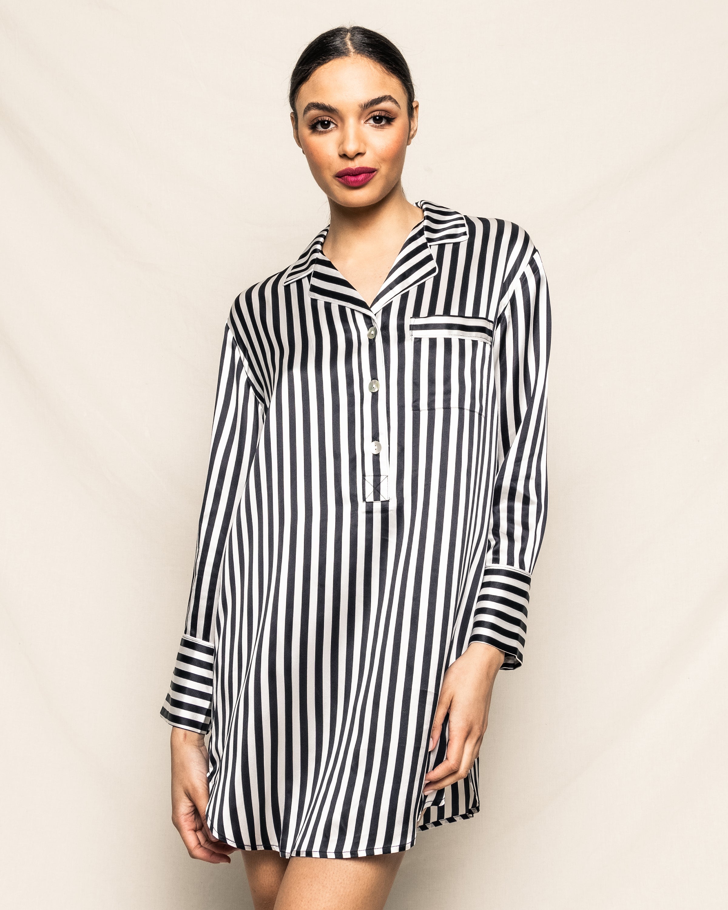 A woman stands against a light beige background, looking at the camera with her hair tied back and bold red lipstick, wearing the Petite Plume Womens Silk Nightshirt in Black and White Bengal Stripe.