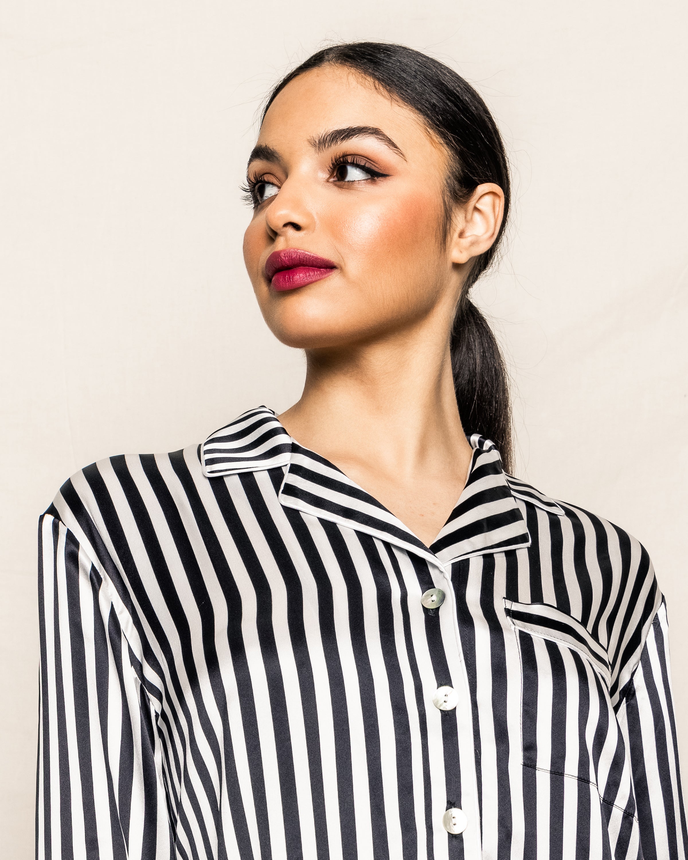 A woman with dark hair in a low ponytail, wearing Petite Plume’s Womens Silk Nightshirt in Black and White Bengal Stripe, looks to her left against a plain light background.