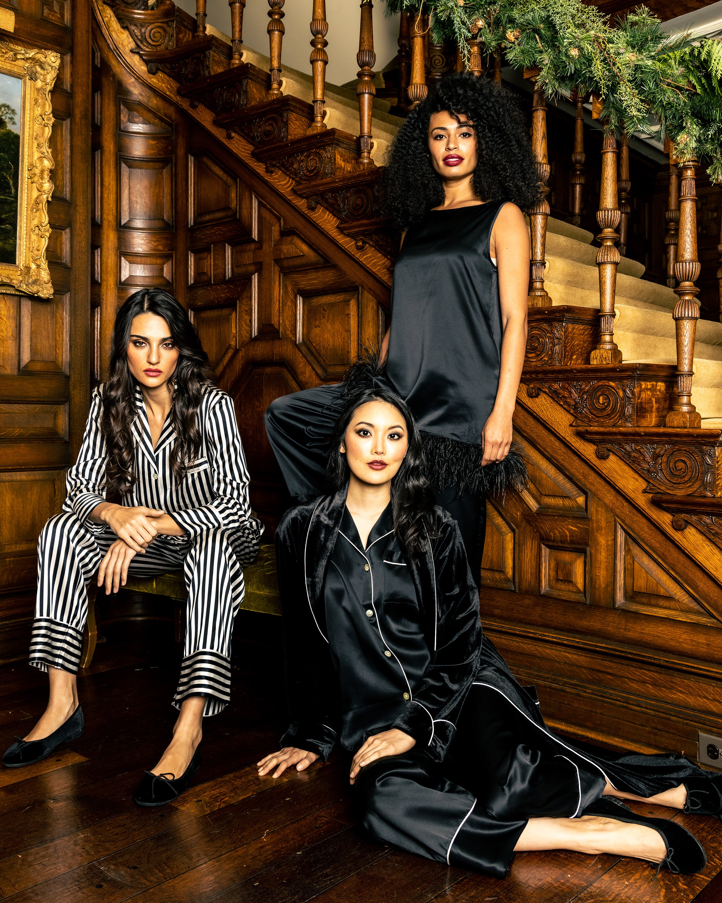 Three women pose on a wooden staircase in luxury sleepwear by Petite Plume. Two sit: one in the Womens Silk Pajama Set in Black and White Bengal Stripe, the other in black, while a third stands behind them in a black satin nightgown with feather trim.