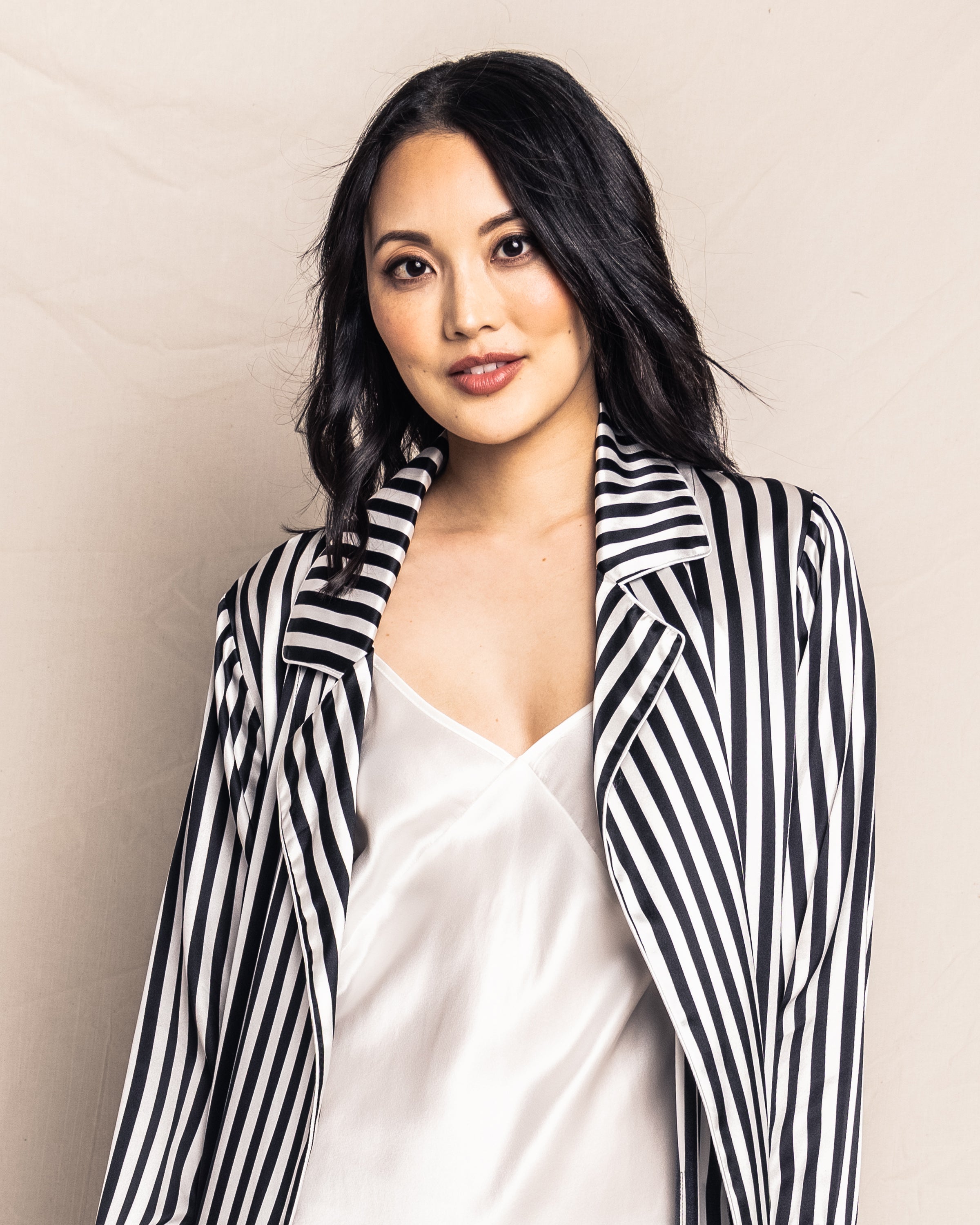 A person with shoulder-length dark hair, wearing Petite Plumes Womens Silk Long Robe in Bengal Stripe over a white top, stands against a neutral background and gazes calmly into the camera.