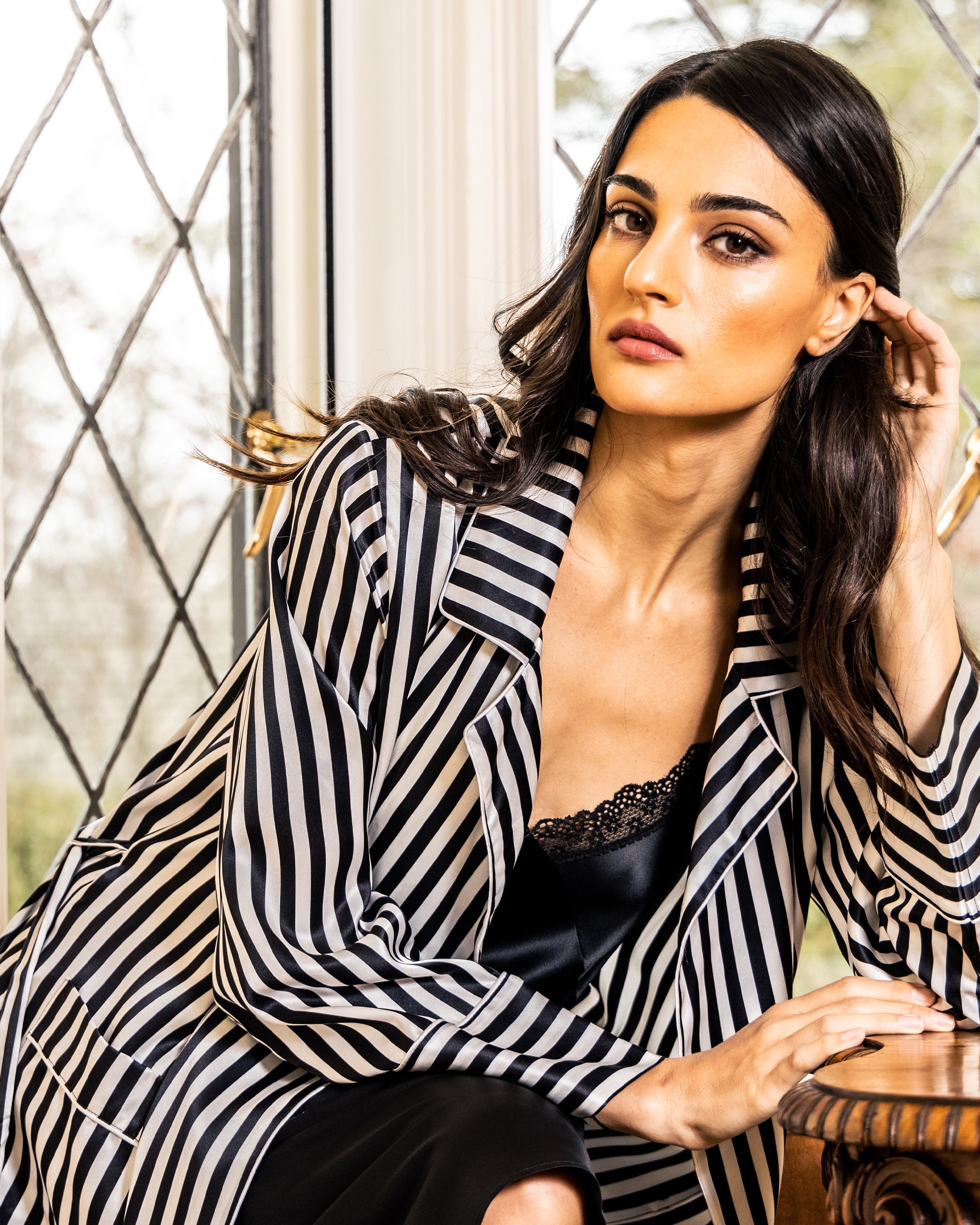 A woman with long dark hair sits indoors by a diamond-paned window, wearing a Bengal stripe silk long robe from Petite Plume over a black lace top, gazing calmly at the camera.