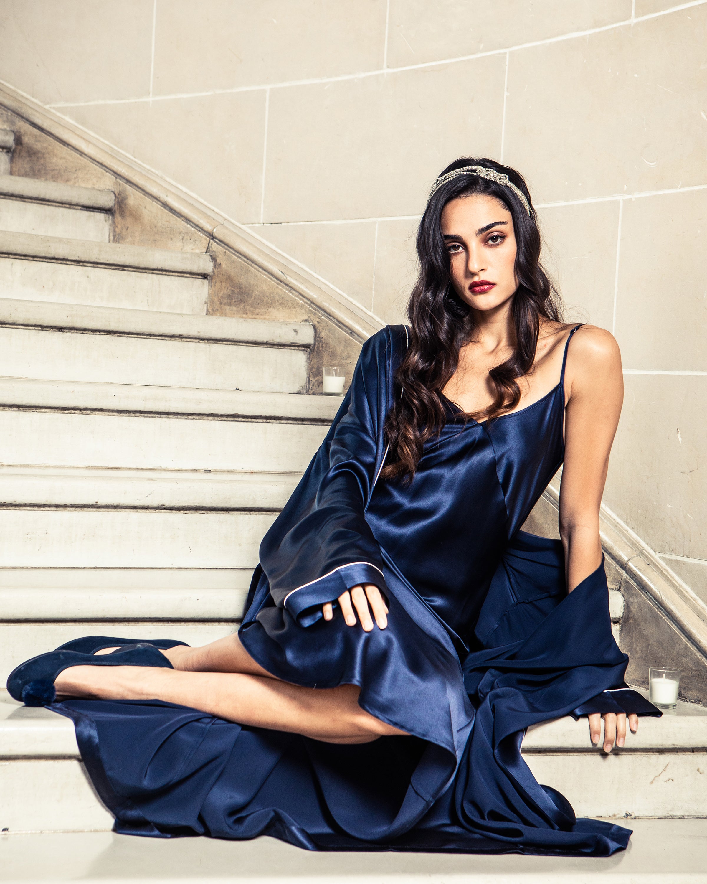 A woman in a Petite Plume Womens Silk Long Robe in Navy, with long, wavy hair and a headband, sits on a stone staircase. Her arm rests on one knee as she gazes at the camera, the luxurious 22mm silk flowing around her under soft lighting.
