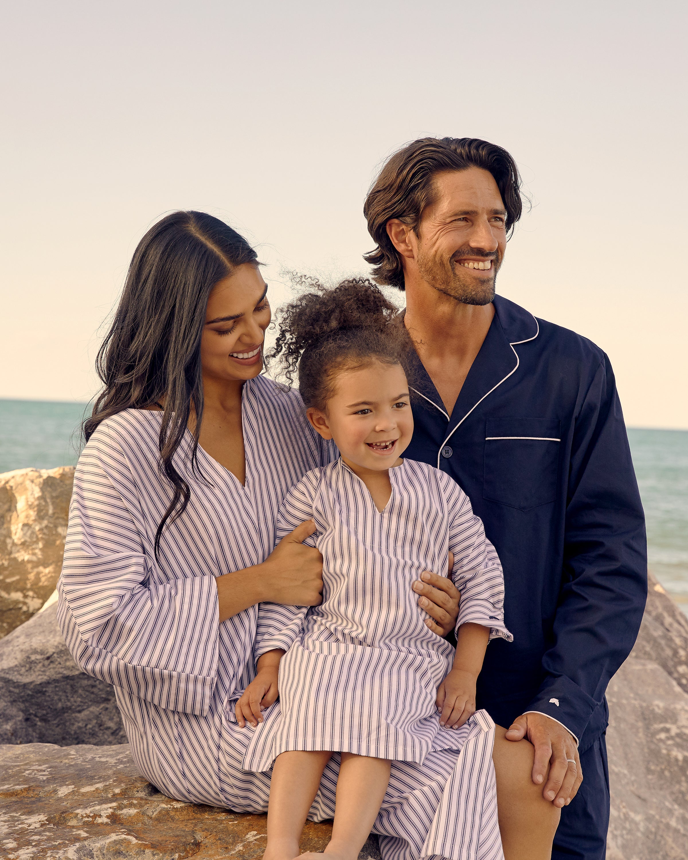 A smiling family sits together on rocks by the ocean. The woman and child wear matching Petite Plume cotton outfits, with the girl in a Navy French Ticking Twill Caftan, while the man sports a dark shirt. The child sits on the womans lap as they gaze into the distance.